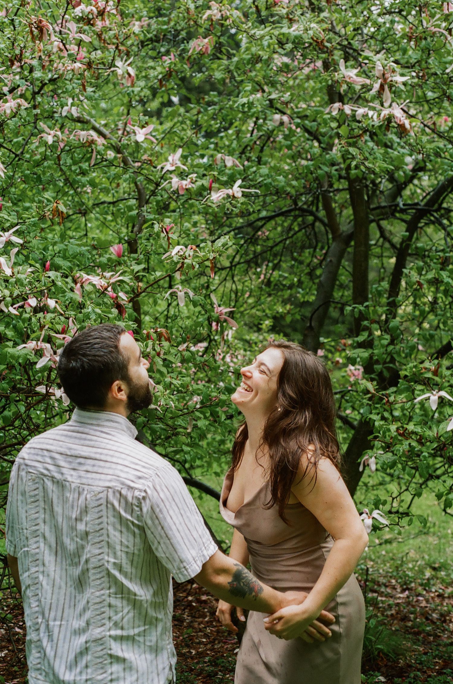 A joyful couple holds hands and laughs together beneath blooming magnolia trees in a lush green garden.