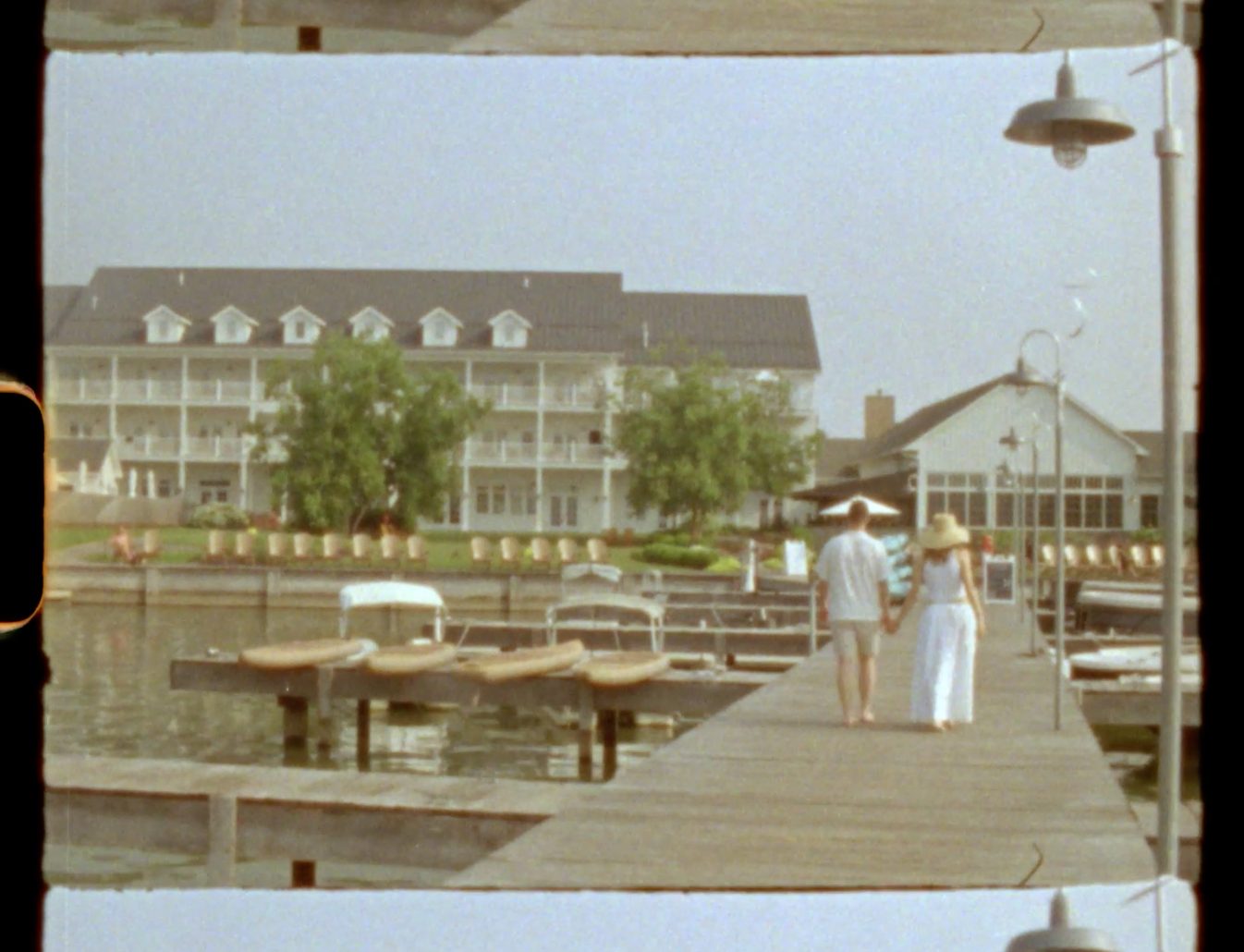Two people stroll along a wooden dock near a large white lakeside resort hotel on a sunny summer day.