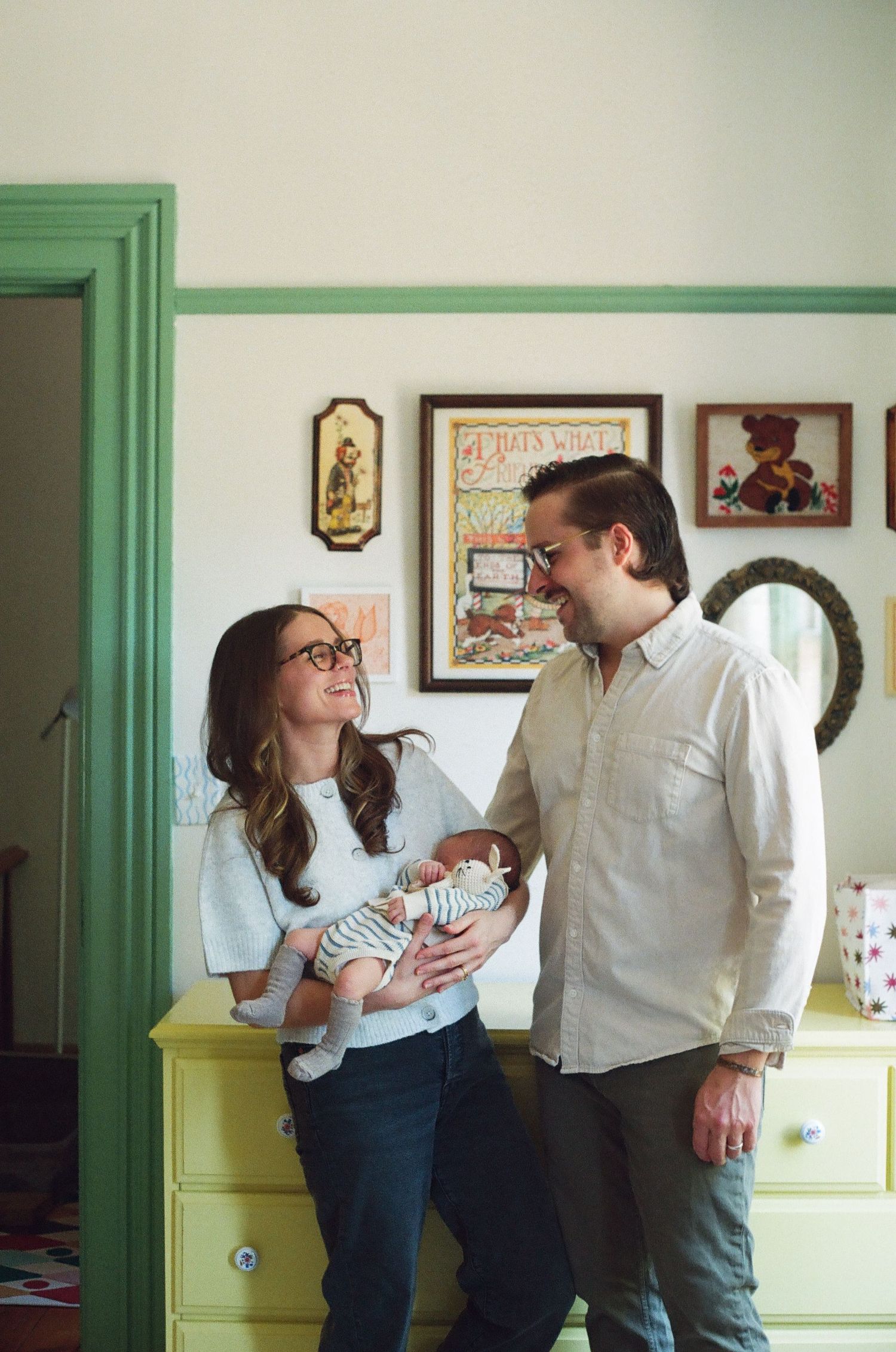 A couple smiles at each other while holding their newborn baby in a cozy, vintage-decorated nursery room.