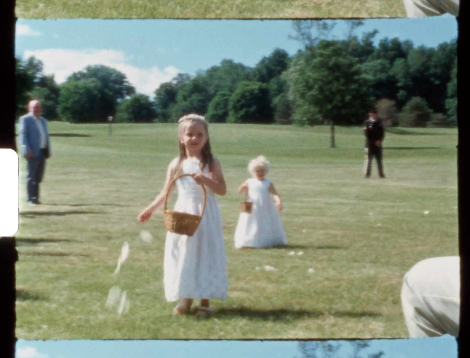 Two young flower girls in white dresses carrying baskets walk across a green lawn during an outdoor wedding ceremony.