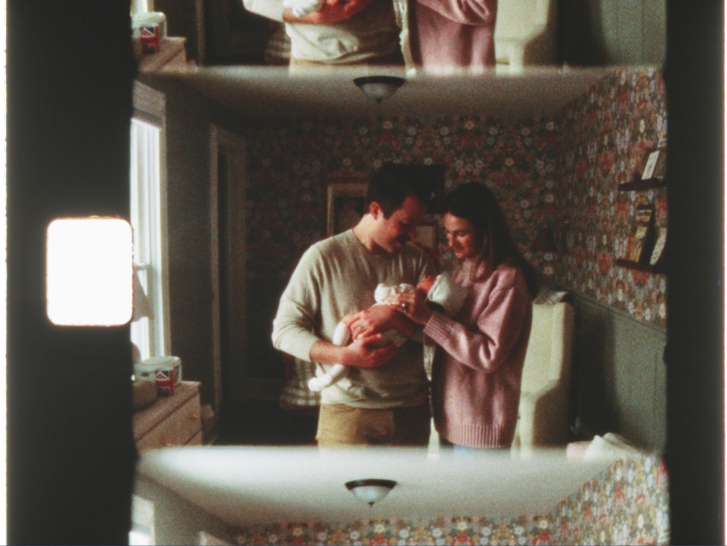 A couple admires their newborn baby in a warmly lit room with floral wallpaper, captured in a vintage film photo.