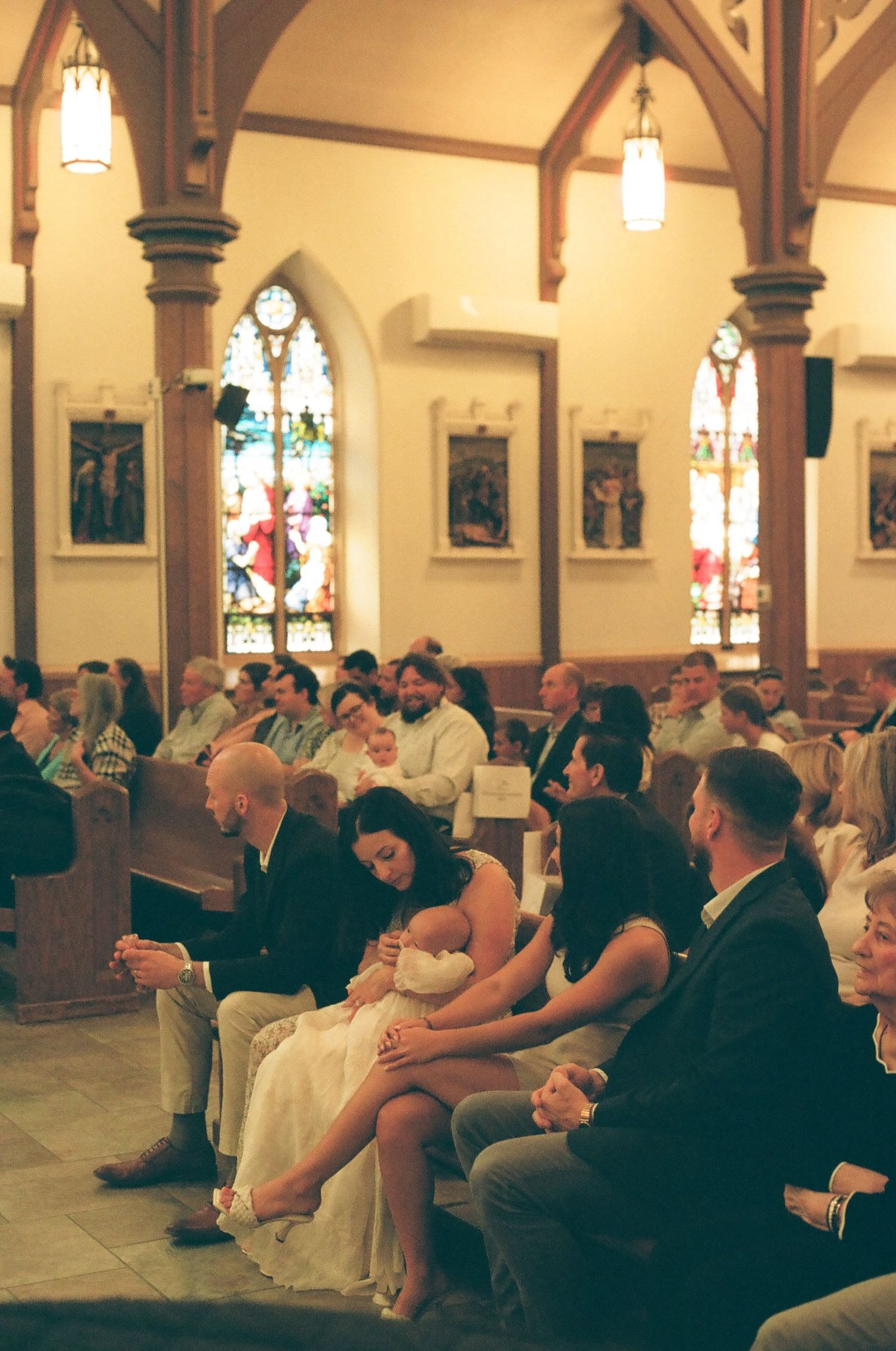 Guests seated in church pews during a wedding ceremony, with colorful stained glass windows in the background.