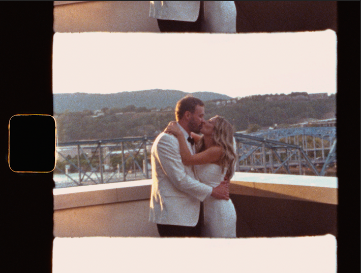 A couple shares a romantic kiss at their wedding on a rooftop venue with scenic hills and a bridge in the background.