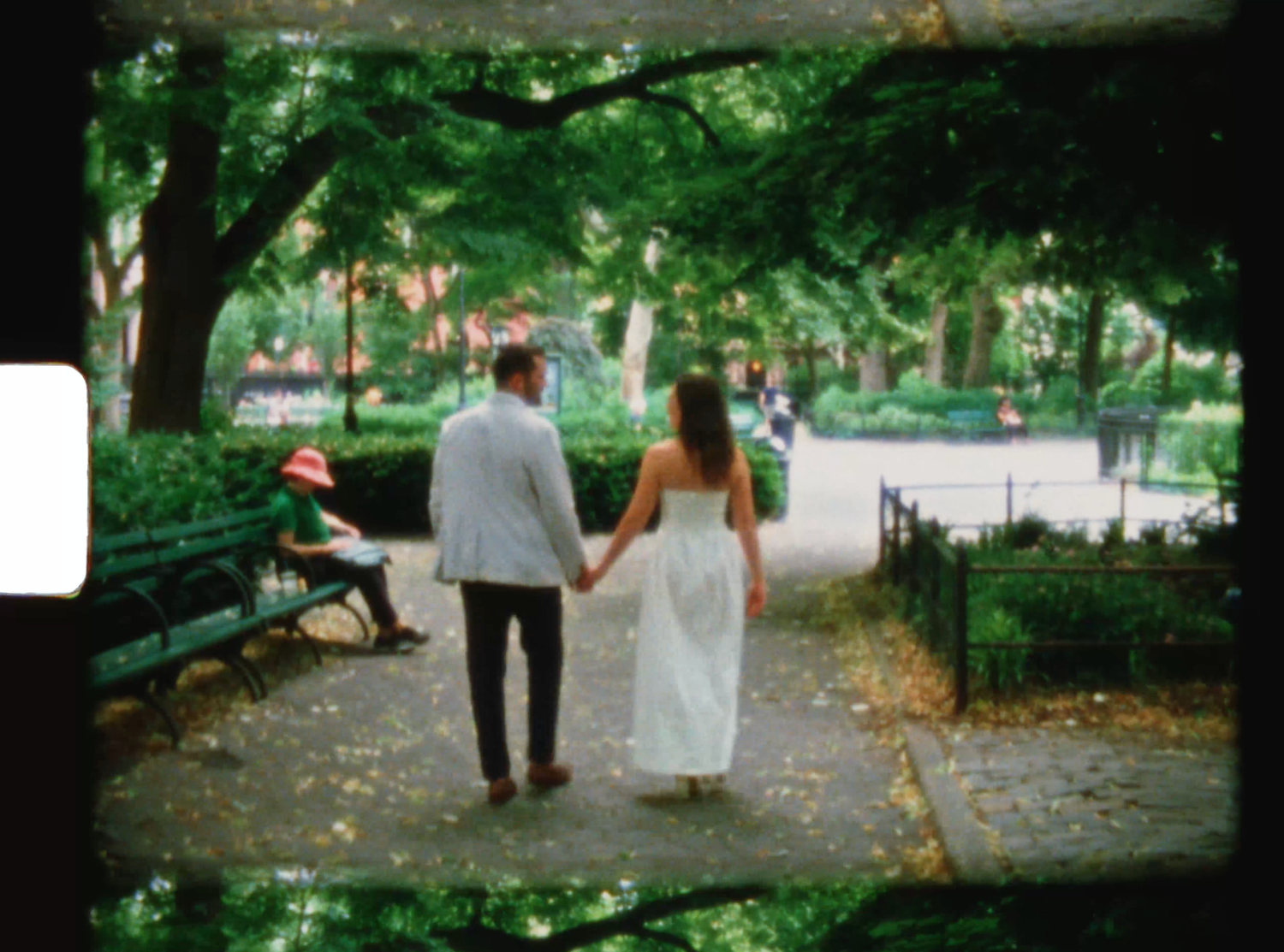 Couple holding hands walking through a lush green park on a tree-lined path, woman in white dress, vintage film photo.