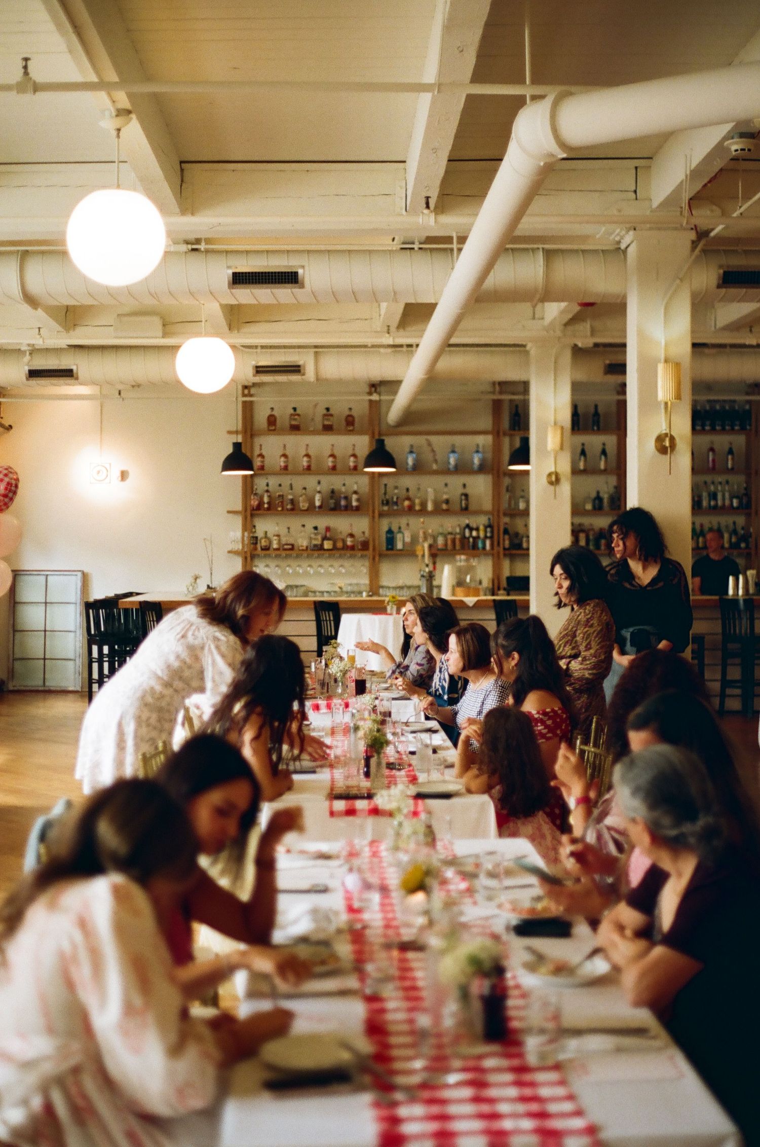Women gathered at a long red checkered table in a warmly lit restaurant with shelves of bottles in background.