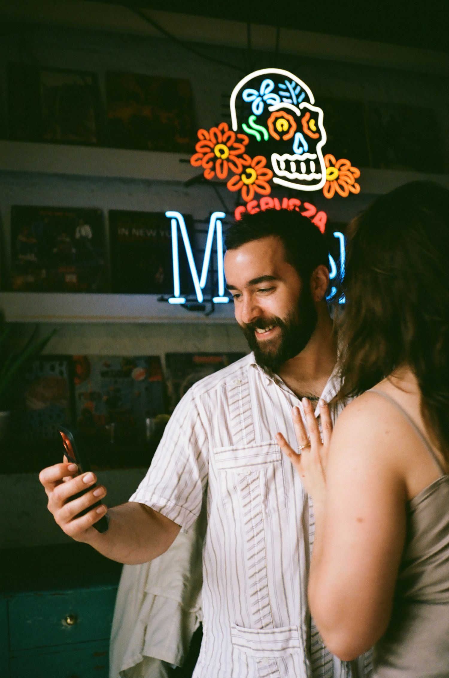 Smiling bearded man holding phone near neon skull sign in colorful bar setting.