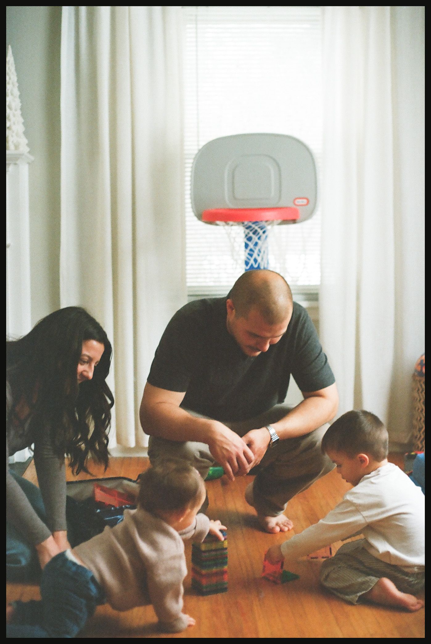 Family with two young children playing on hardwood floor indoors, with a small basketball hoop mounted on the wall behind them.