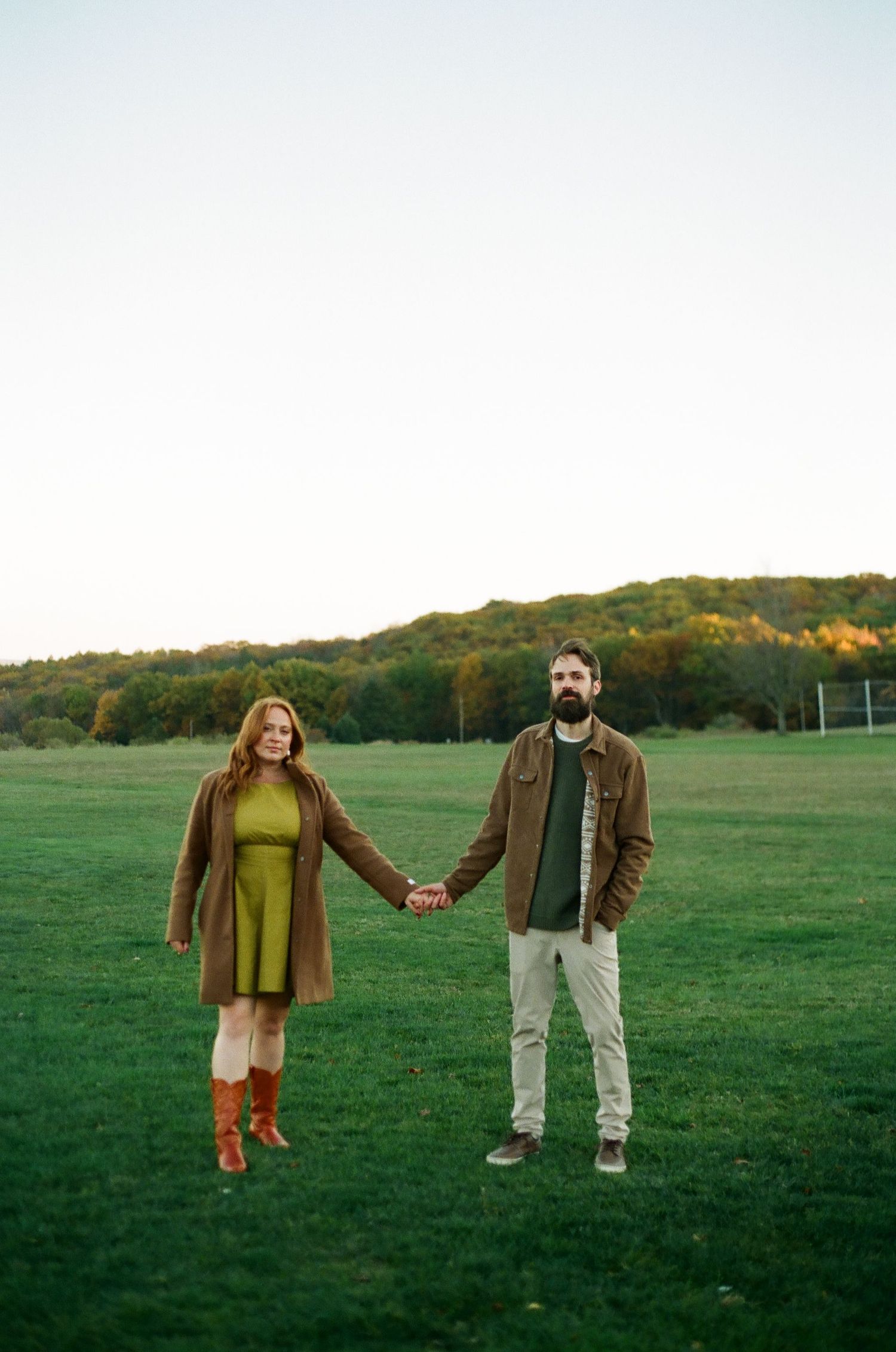 Couple holding hands in green field with autumn trees, woman in olive dress and brown boots, man in brown jacket.