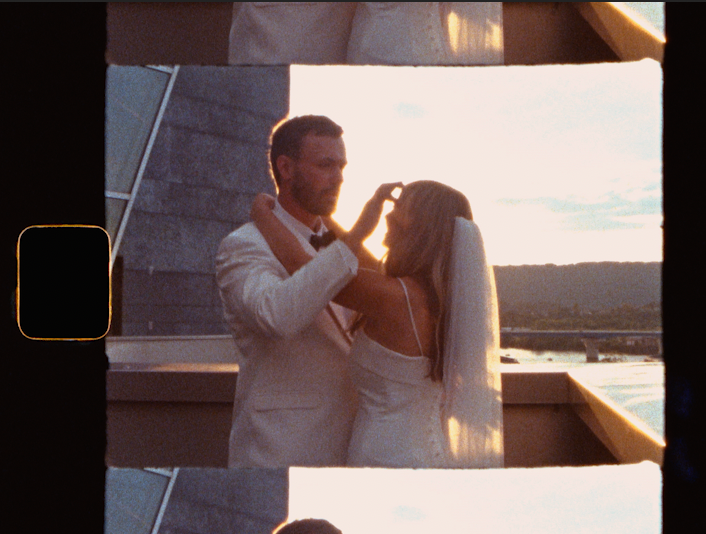 A bride and groom share a romantic moment at sunset on a rooftop, the groom gently touching the bride's hair.