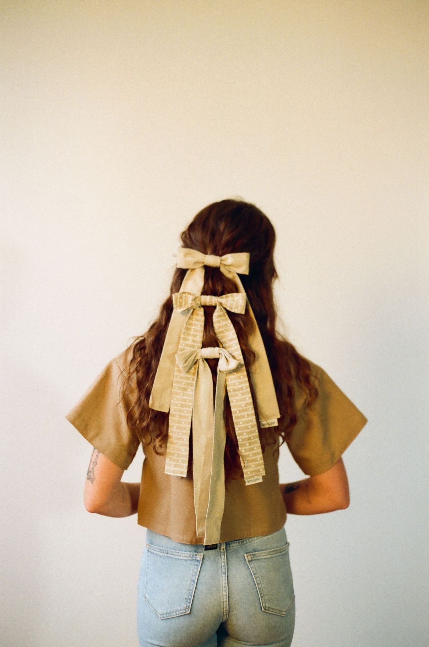 Woman with brown hair styled with three cream ribbon bows, wearing a tan crop top and light blue jeans, viewed from behind.
