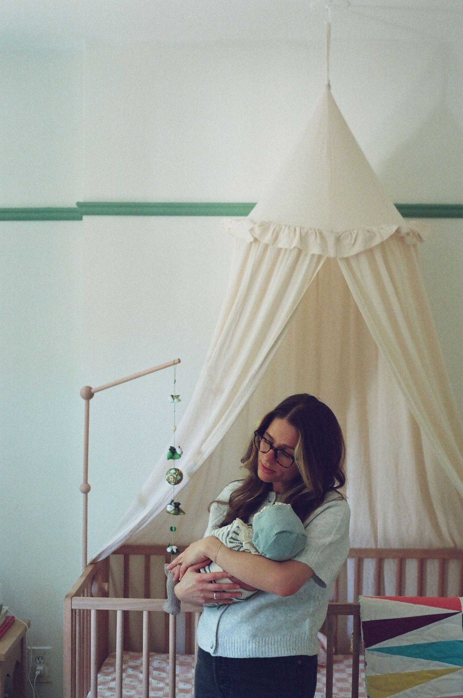 Mother holding newborn baby in nursery with white canopy crib and colorful mobile.