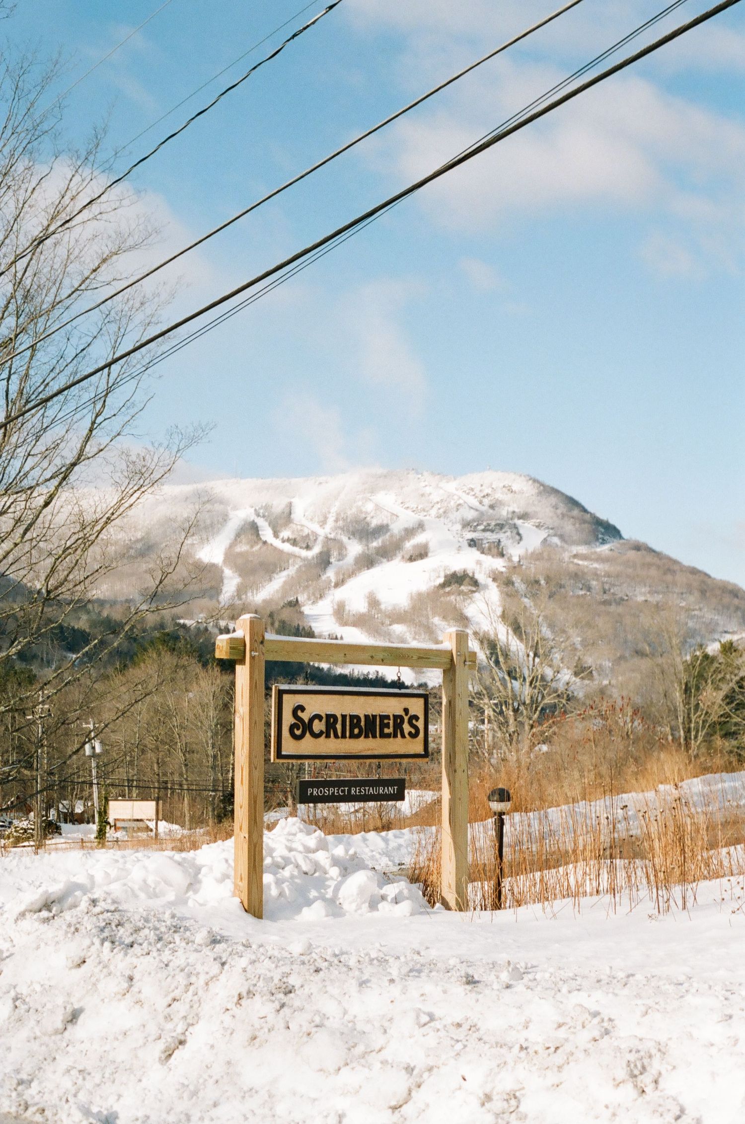 Scribner's restaurant sign in winter snow with ski mountain slopes visible in the background under a blue sky.