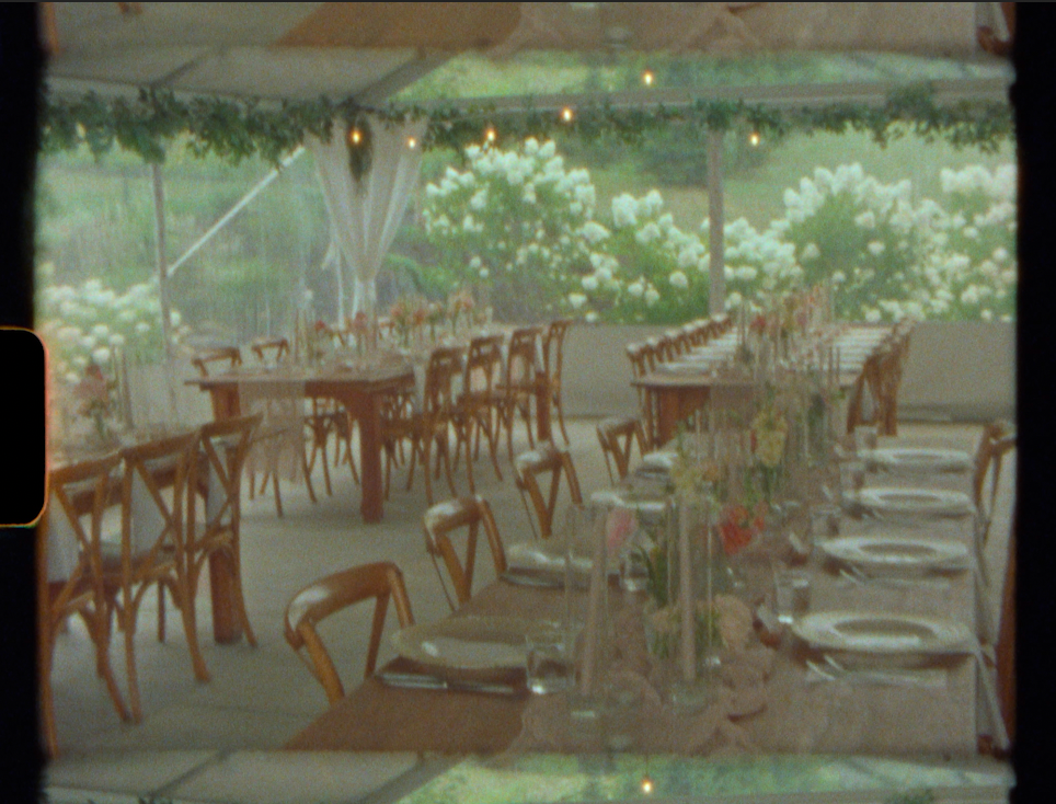 Elegant outdoor wedding reception under a tent with wooden cross-back chairs, long dining tables, and lush white floral greenery backdrop.