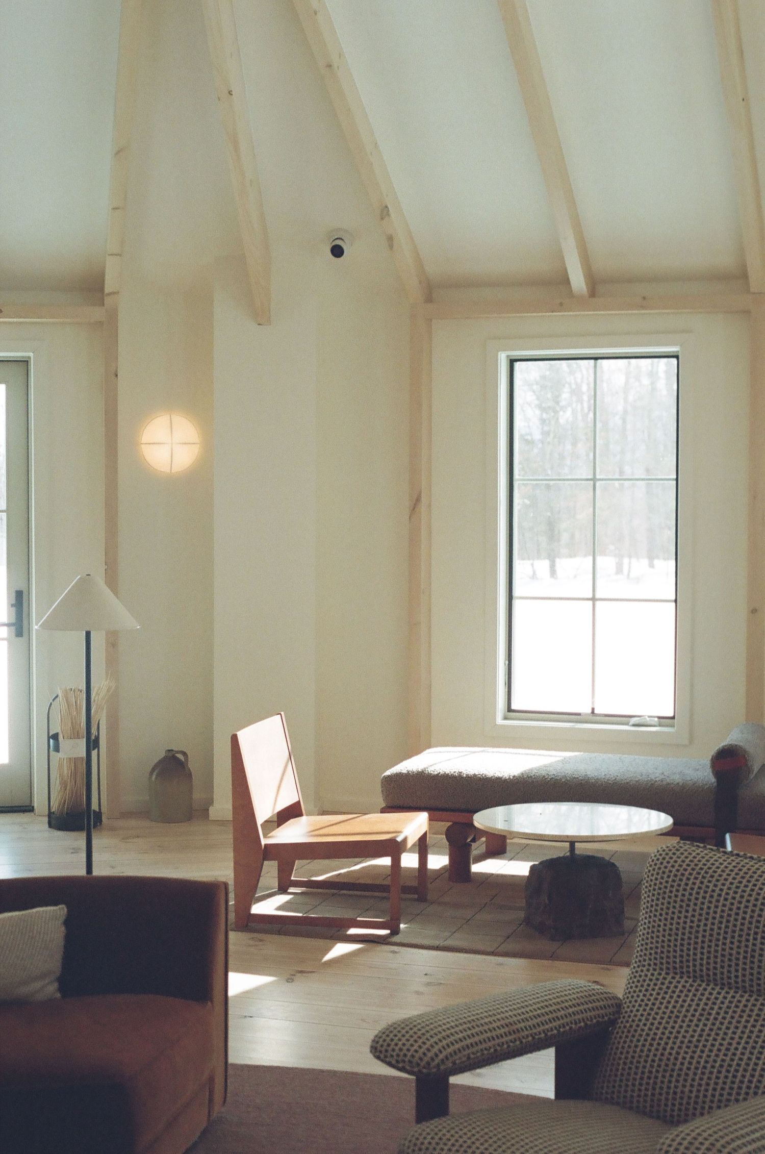 Minimalist barn-style living room with vaulted white ceiling, wooden chairs, and houndstooth seating near a large window.