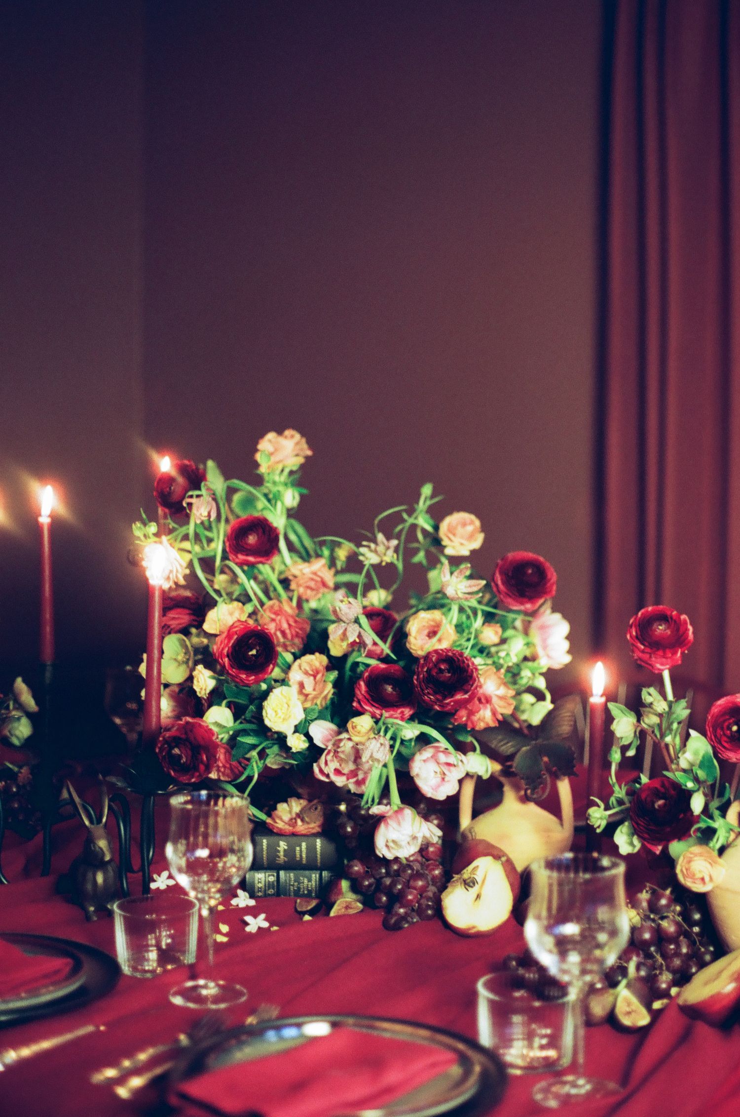 Elegant candlelit tablescape with deep red floral centerpiece, ranunculus blooms, tapered candles, and crystal glassware on crimson linen.