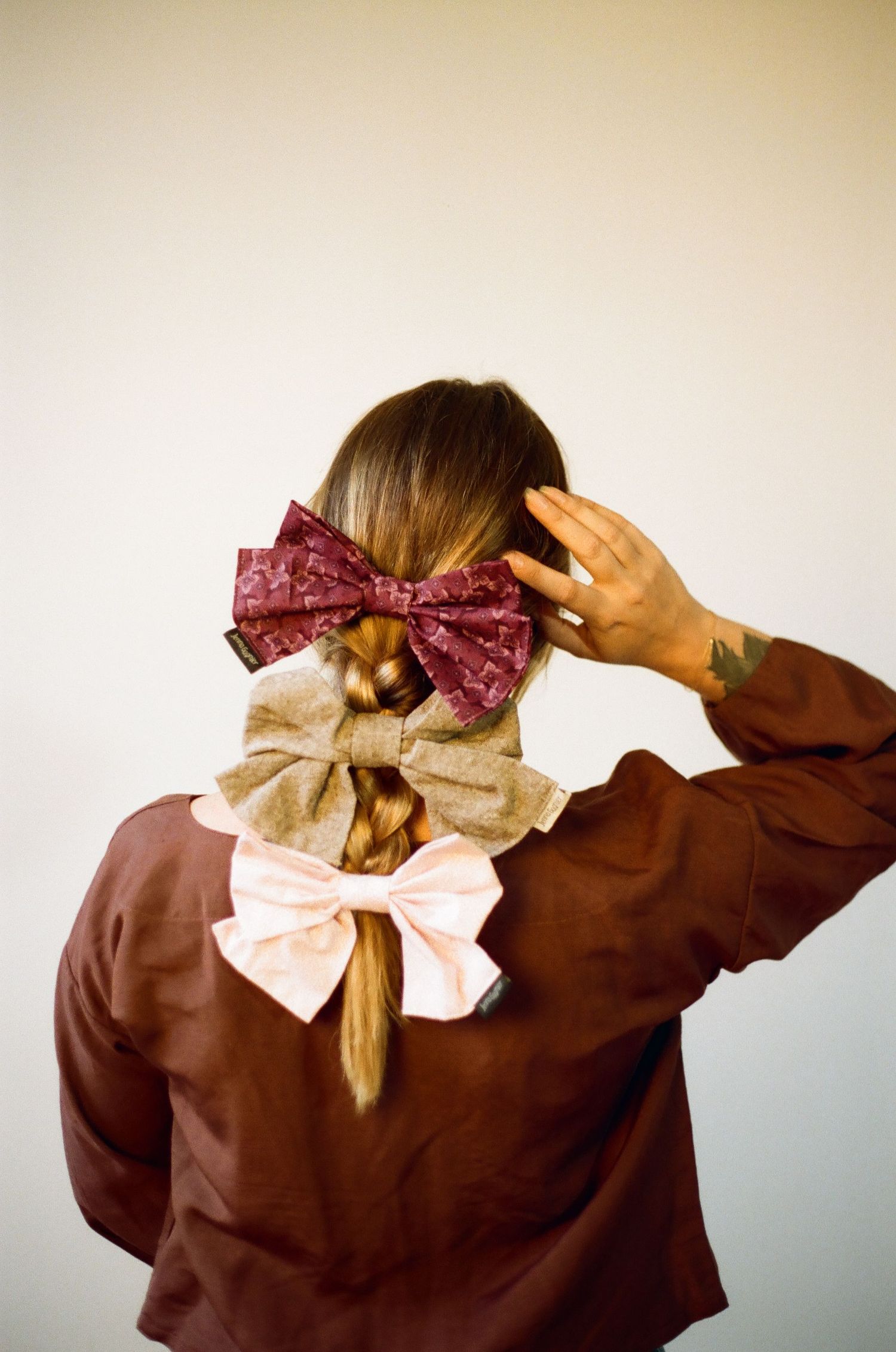 Woman with braided hair styled with three decorative bows in burgundy, tan, and pink, wearing a brown jacket.