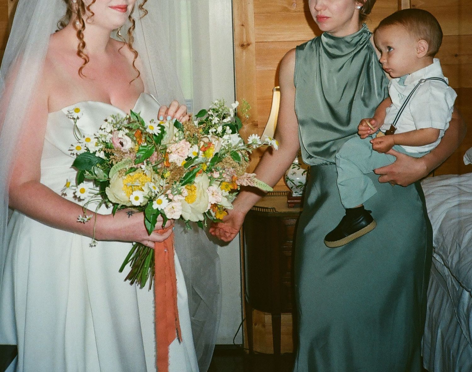 Bride in white gown holding wildflower bouquet stands beside bridesmaid in green dress holding a young child at wedding.