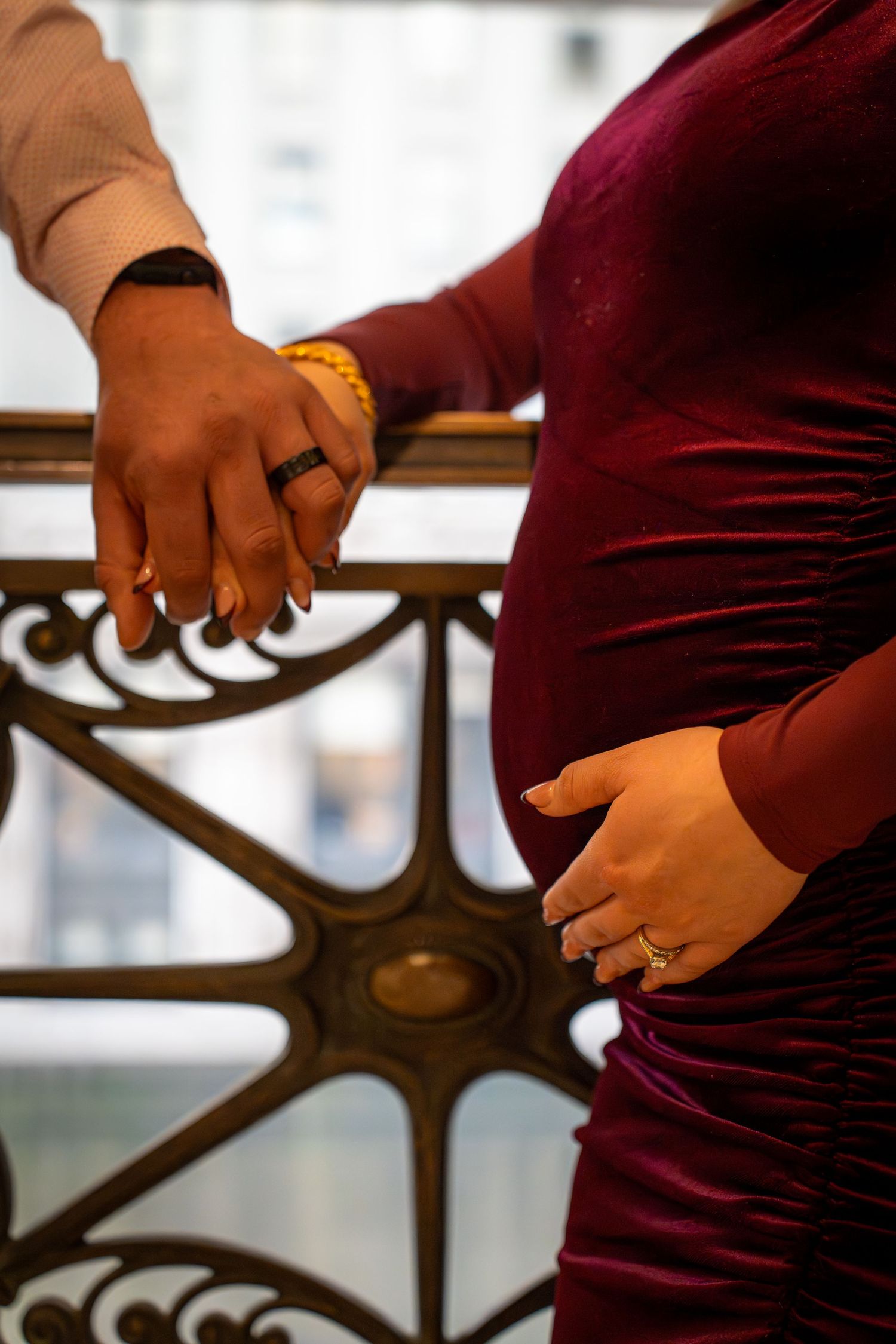 Close-up of hands wearing engagement rings against ornate metalwork railing, with burgundy and brown clothing details.