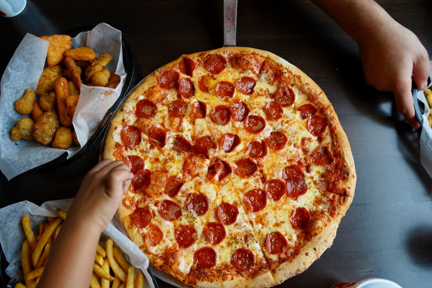 Overhead view of pepperoni pizza and french fries on a dark table.
