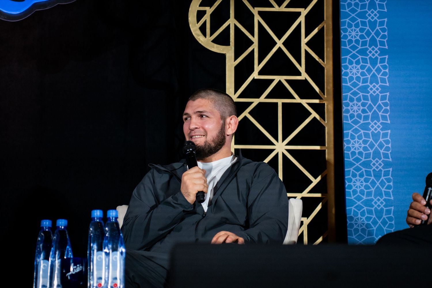 A speaker sits at a panel discussion table with water bottles and decorative gold patterns on a blue wall behind them.