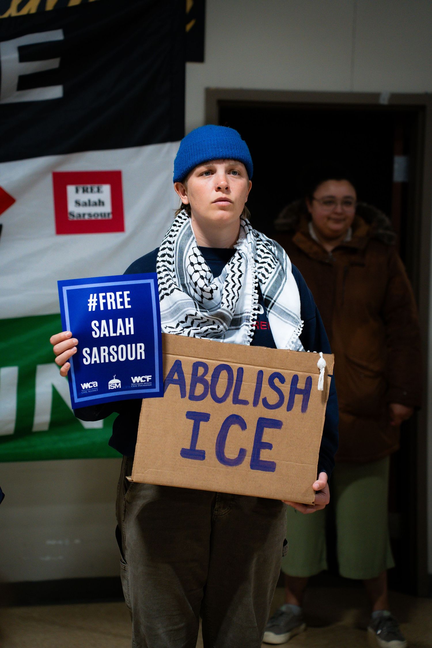 Activist holding Abolish ICE sign and Free Salah Sarsour placard at protest, wearing blue beanie and keffiyeh.