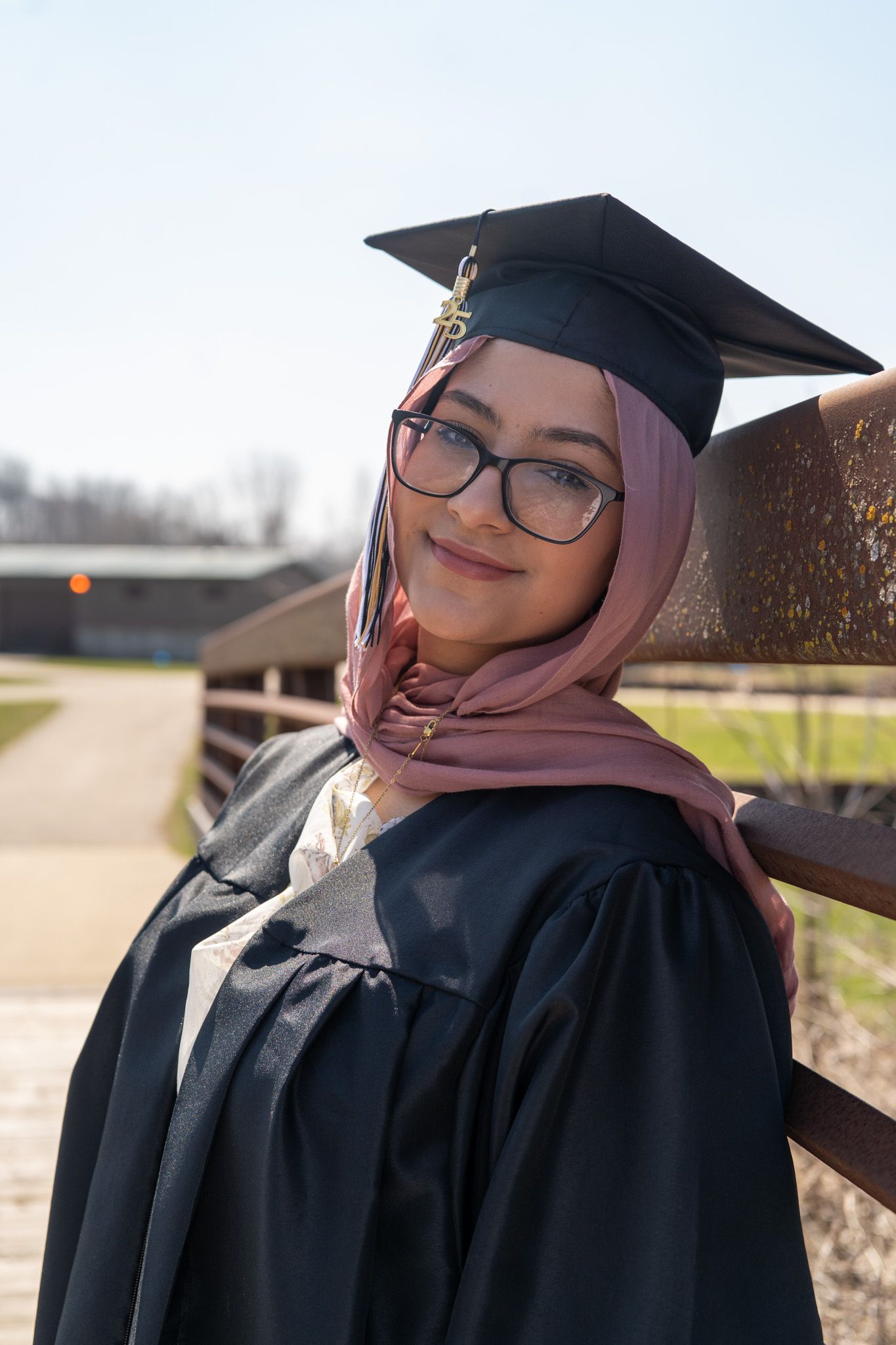 Graduate in black cap and gown poses outdoors on sunny day.