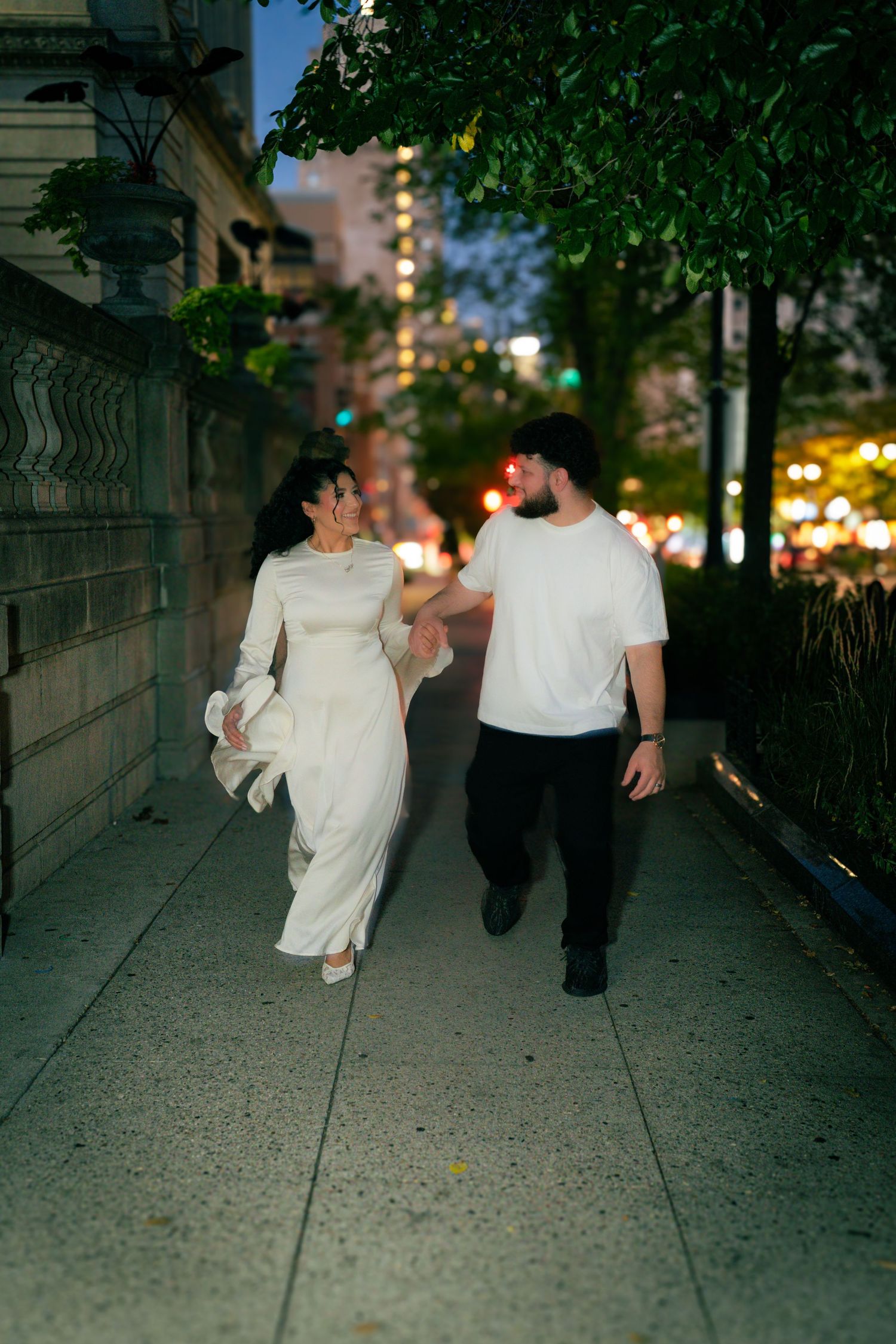 A couple walking together at night on a city sidewalk with trees and urban lighting in the background.