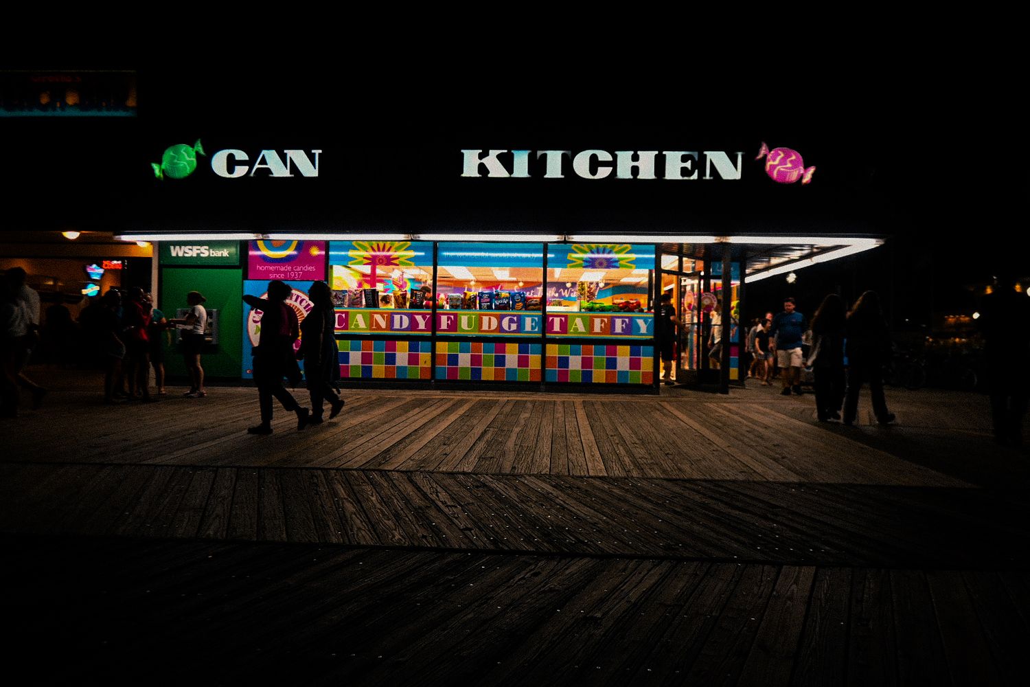 Illuminated food stand with 'Kitchen' sign glowing at night on a wooden boardwalk.