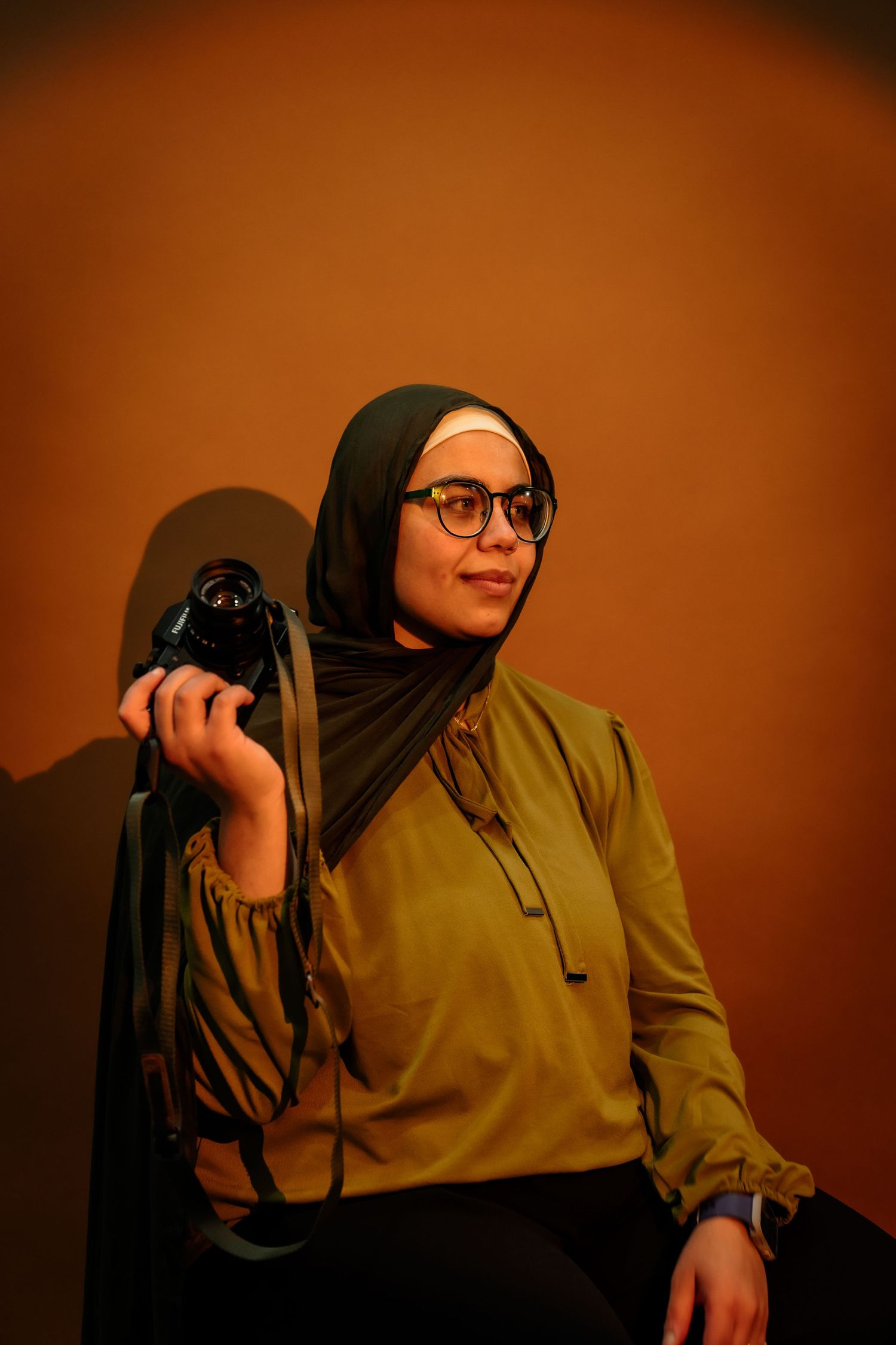 A photographer in a hijab and olive jacket holds a camera while posing against a dark reddish-brown background.