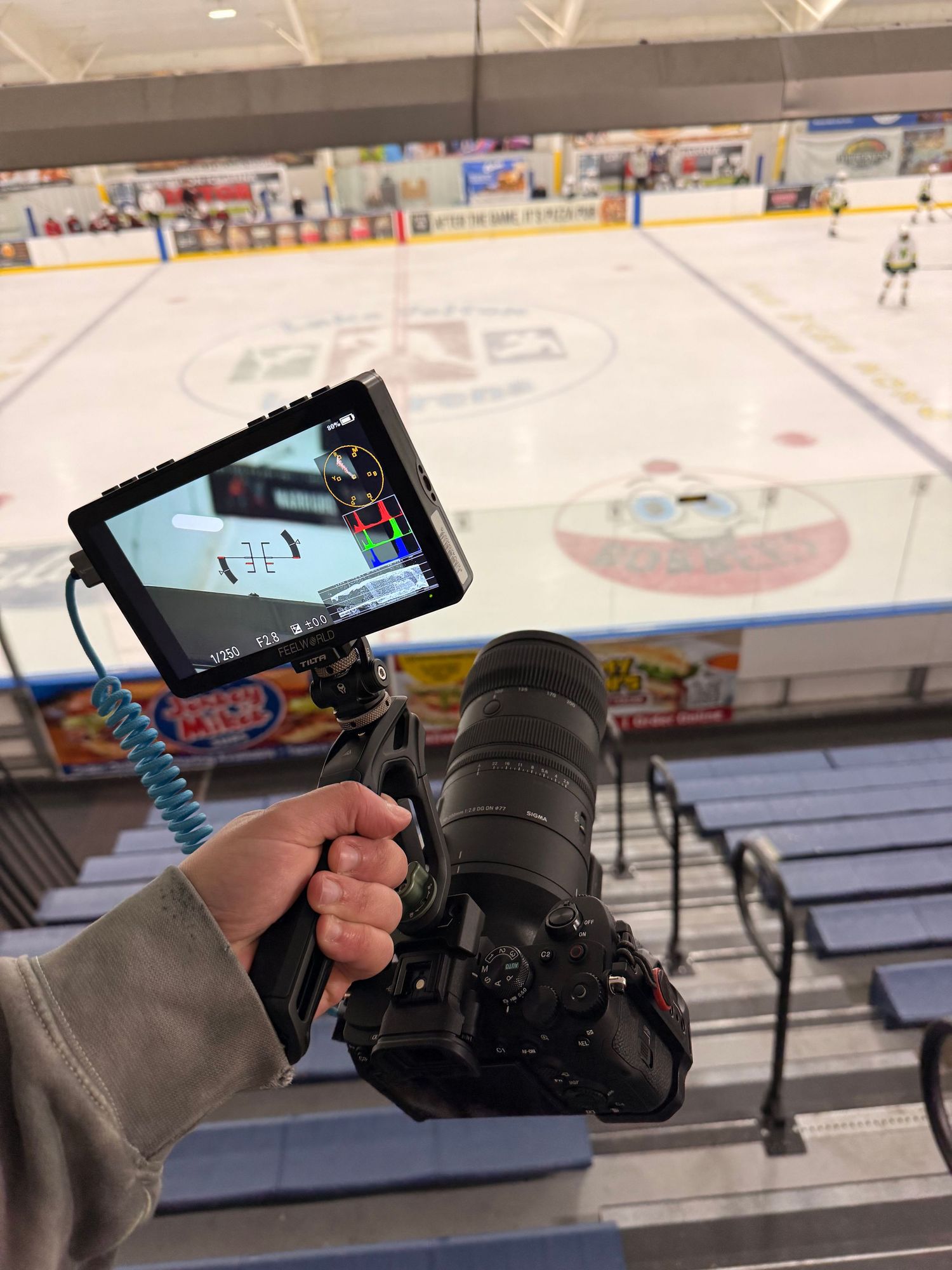 A photographer holds a DSLR camera with an external monitor filming a hockey rink during a game.
