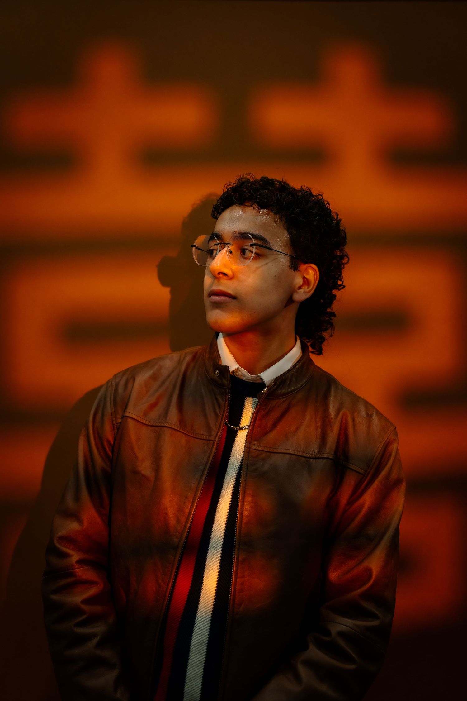 A person in a leather jacket and white tie poses against a dramatic orange sunset background in a moody portrait series.