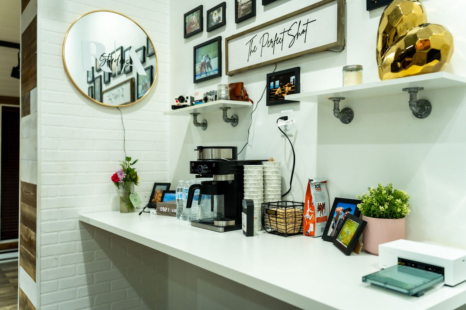 A coffee station with espresso machine and supplies mounted on white brick wall with framed artwork and shelving.