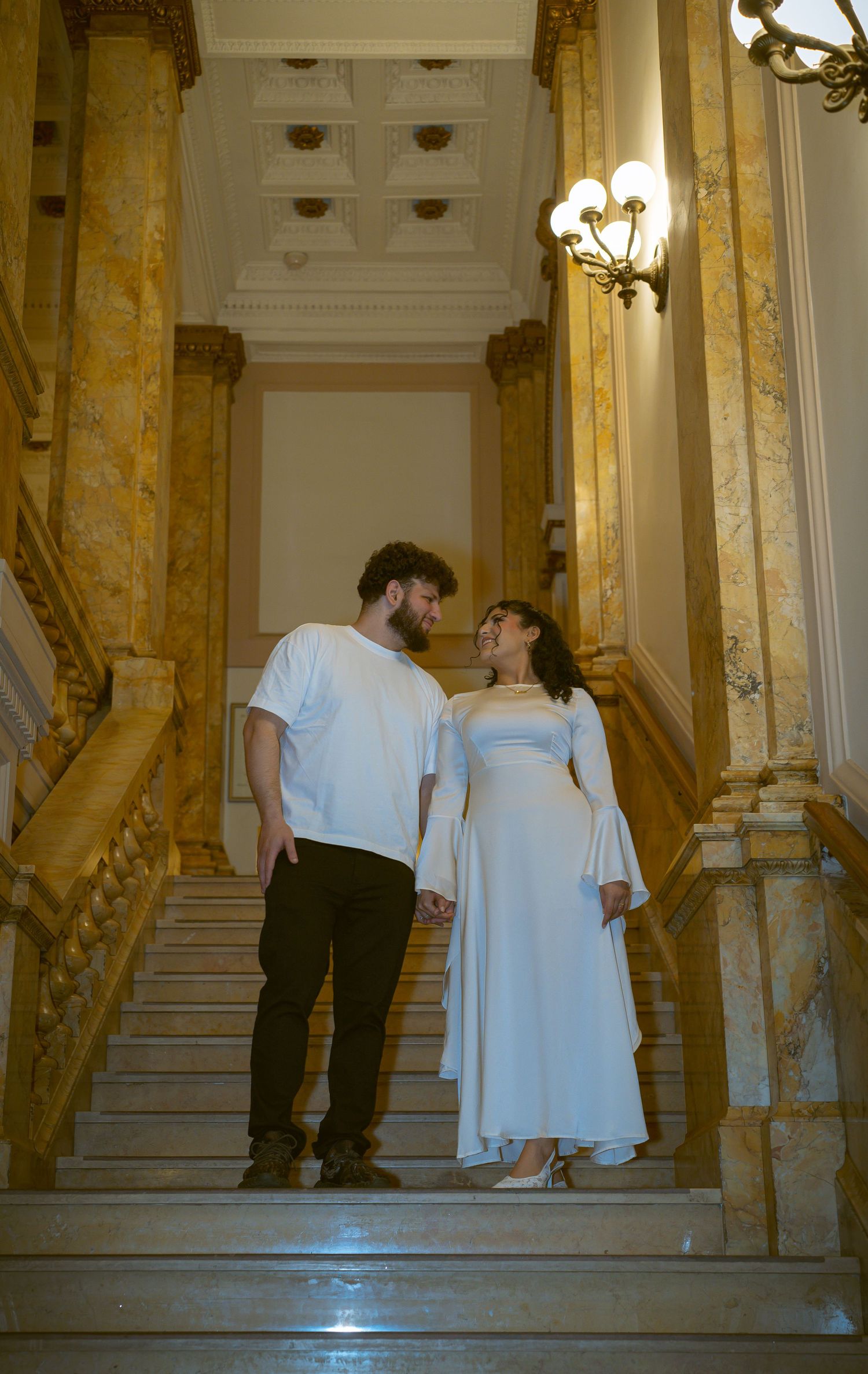 Couple in white attire posing on grand marble staircase.
