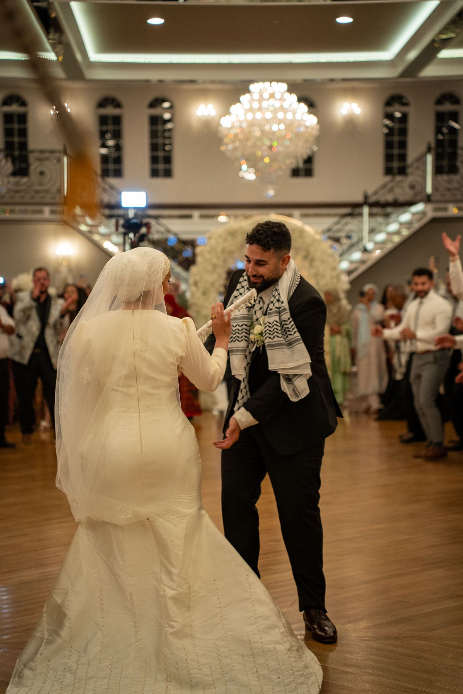 A couple performing a traditional dance in an elegant ballroom with chandeliers overhead.