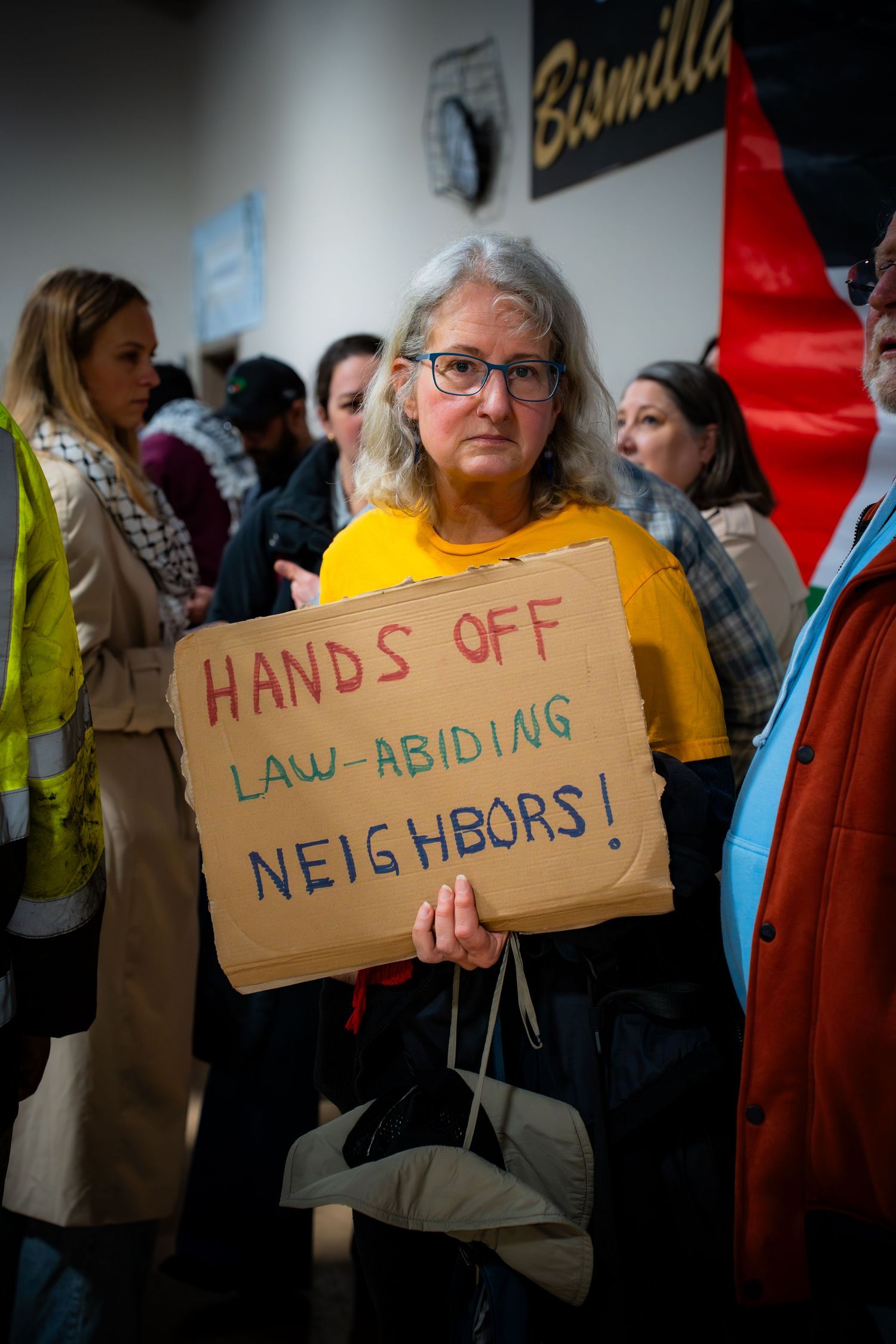 Woman holding Hands Off Law-Abiding Neighbors cardboard sign at indoor immigration protest rally.