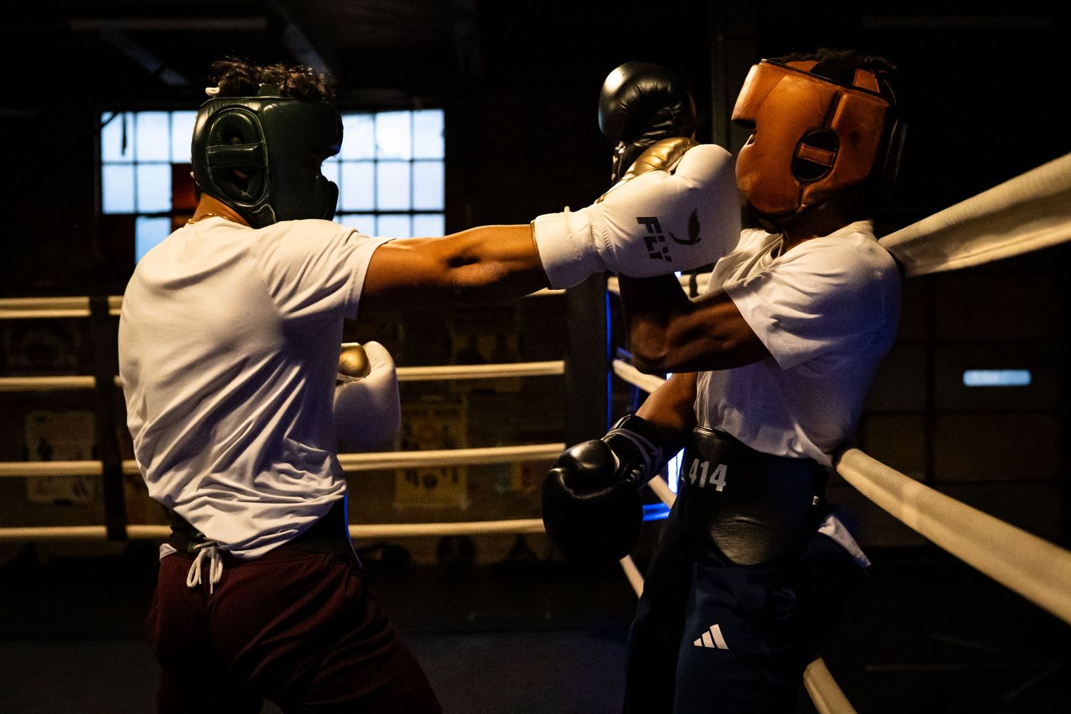 Two boxers spar in a dimly lit boxing ring with ropes and windows visible in the background.