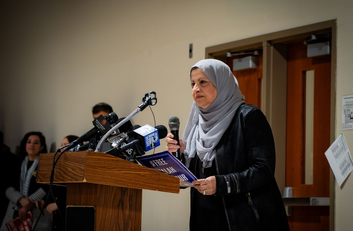 Woman in gray hijab and black jacket speaks at podium with microphones during a press conference.