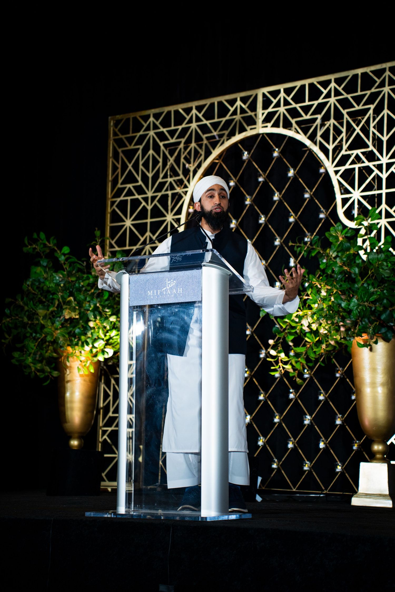 Speaker presenting from a podium on stage with decorative lattice backdrop and potted plants.