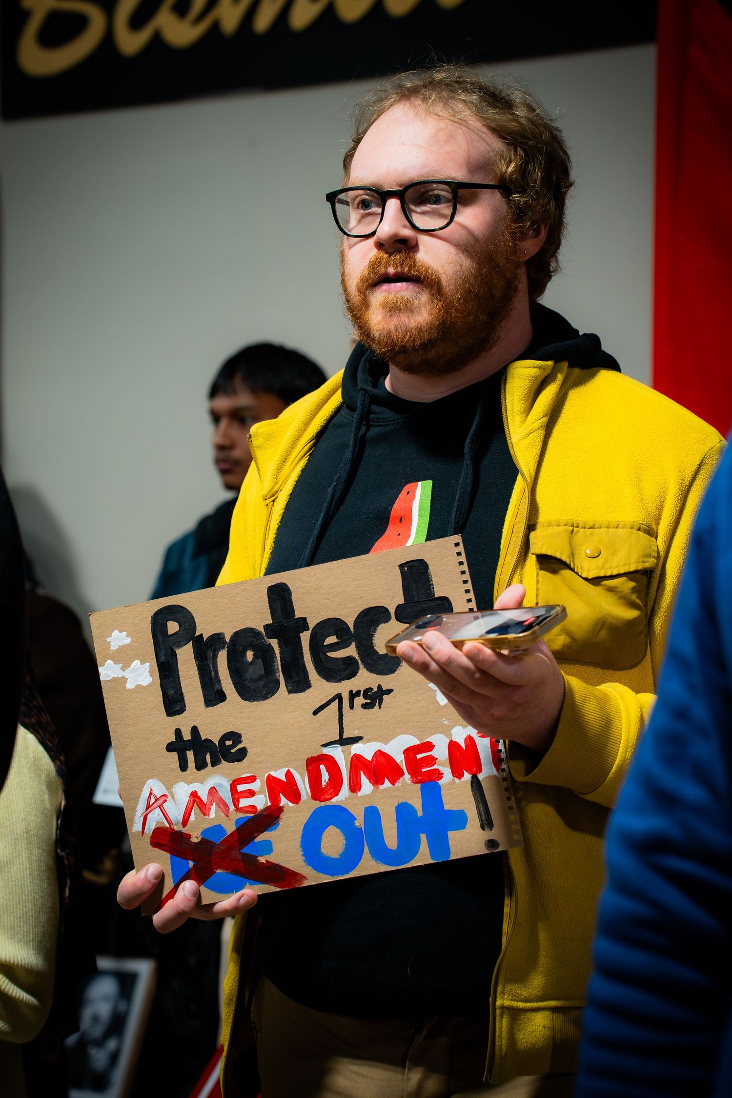 Bearded man in yellow jacket holds cardboard sign reading Protect the 1st Amendment at rally.