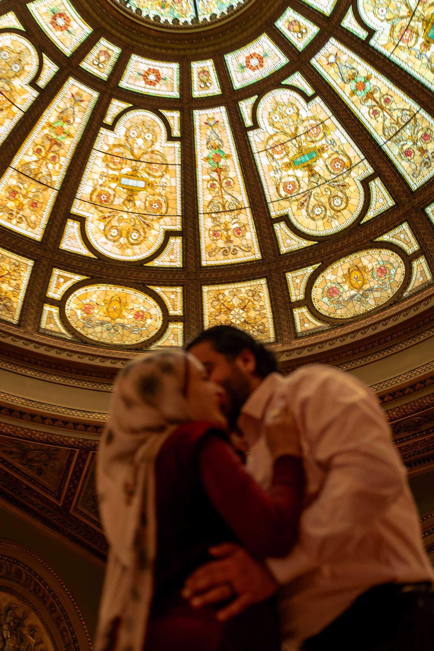 A romantic moment beneath an ornate stained glass dome ceiling with intricate floral and geometric patterns.