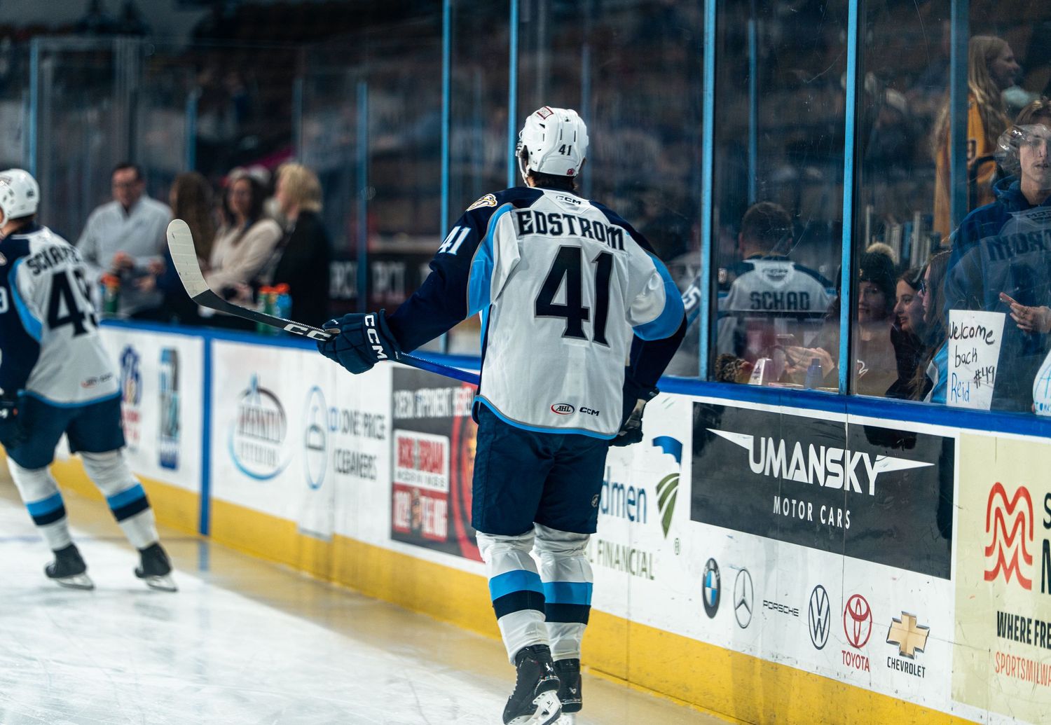 Ice hockey players skate across a blue-tinted rink during a competitive match with spectators in the background.