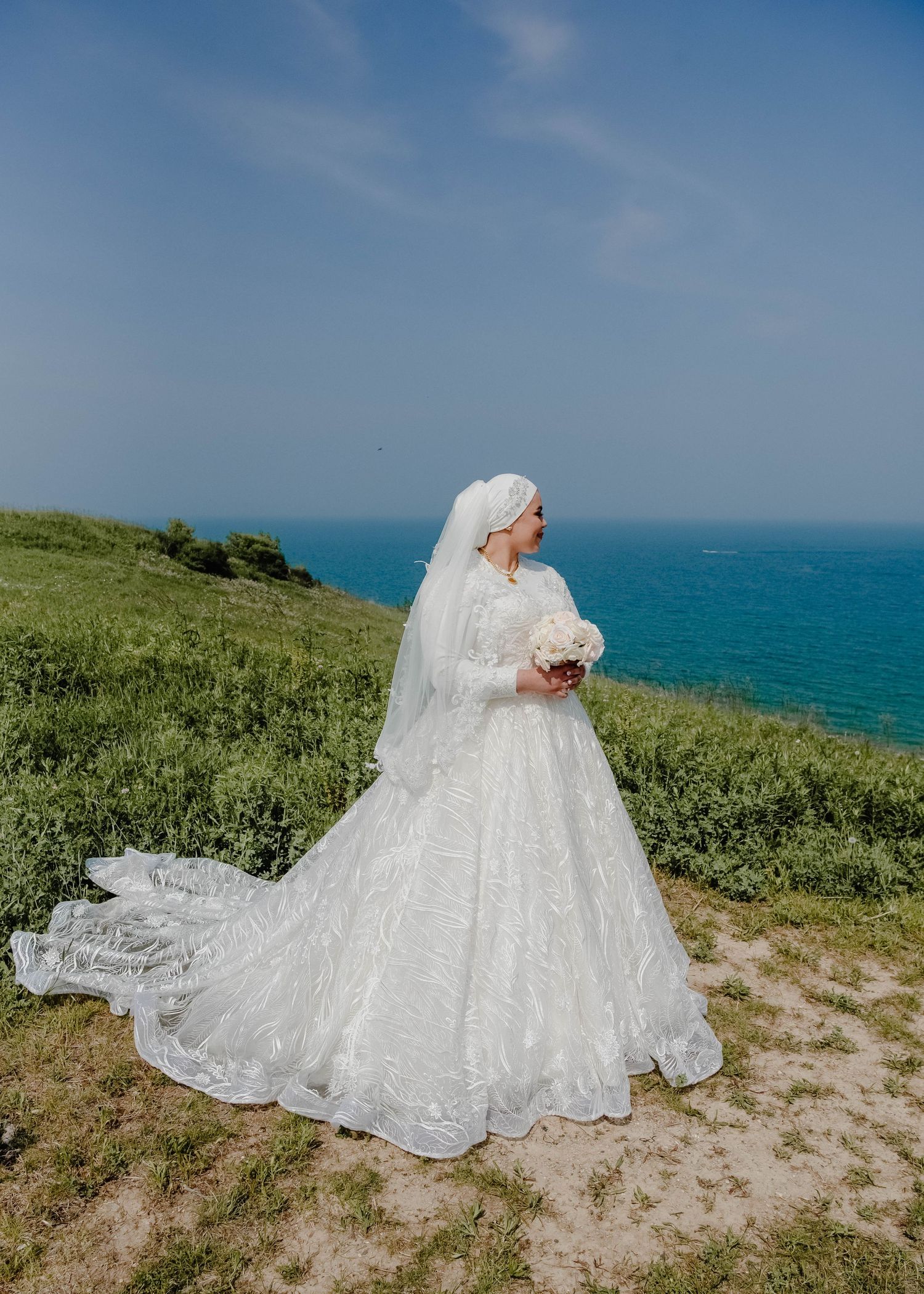 White wedding dress flowing in breeze against ocean backdrop on grassy cliff.