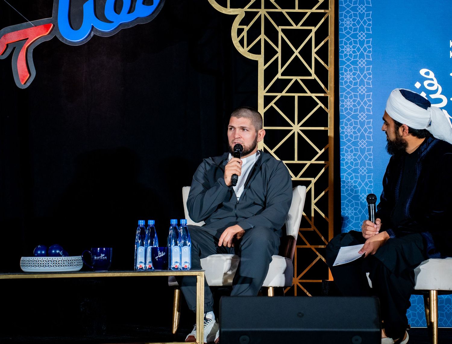 Panel discussion setup on stage with water bottles and chairs against decorative backdrop.