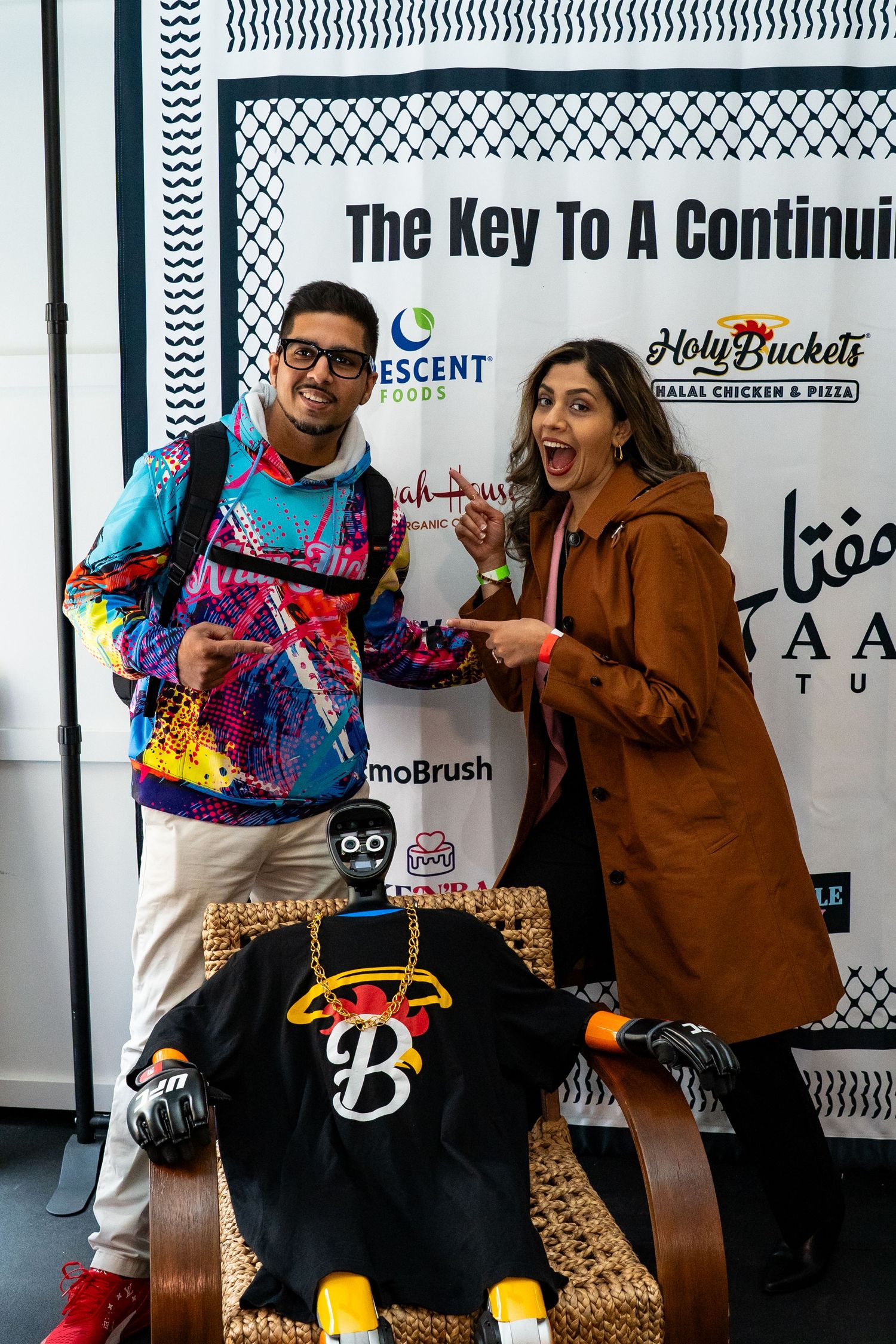 Two people pose excitedly in front of a promotional backdrop with a skateboard at a branded event.