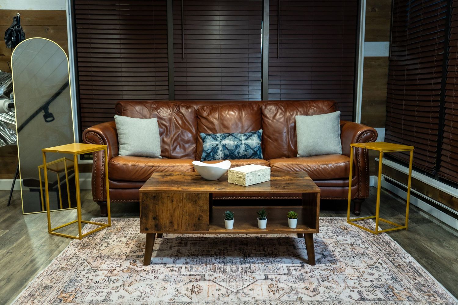 Modern living room with brown leather couch, wooden coffee table, gold side tables and patterned area rug.