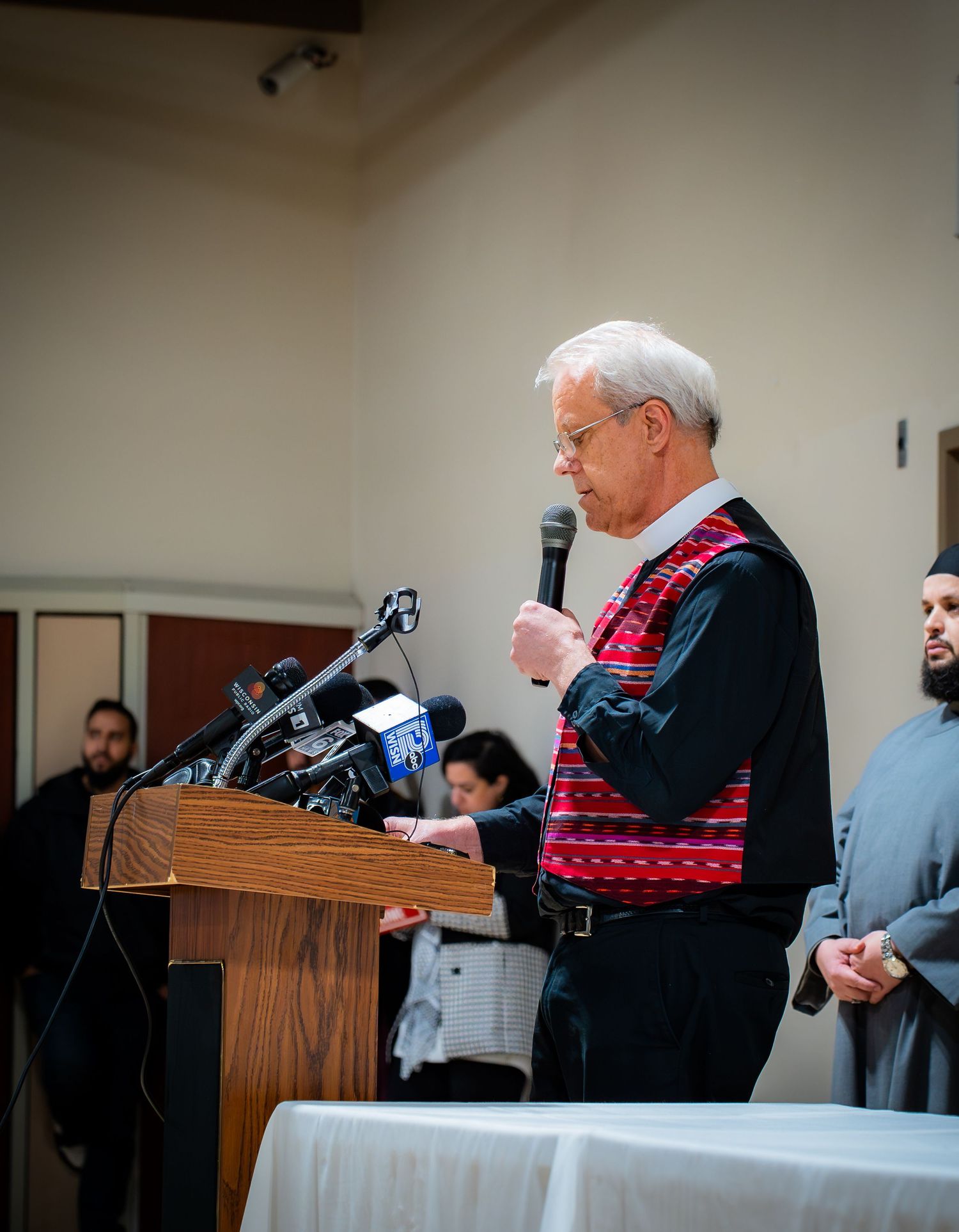 Man in traditional vest speaks at podium with microphones at an interfaith community press conference.