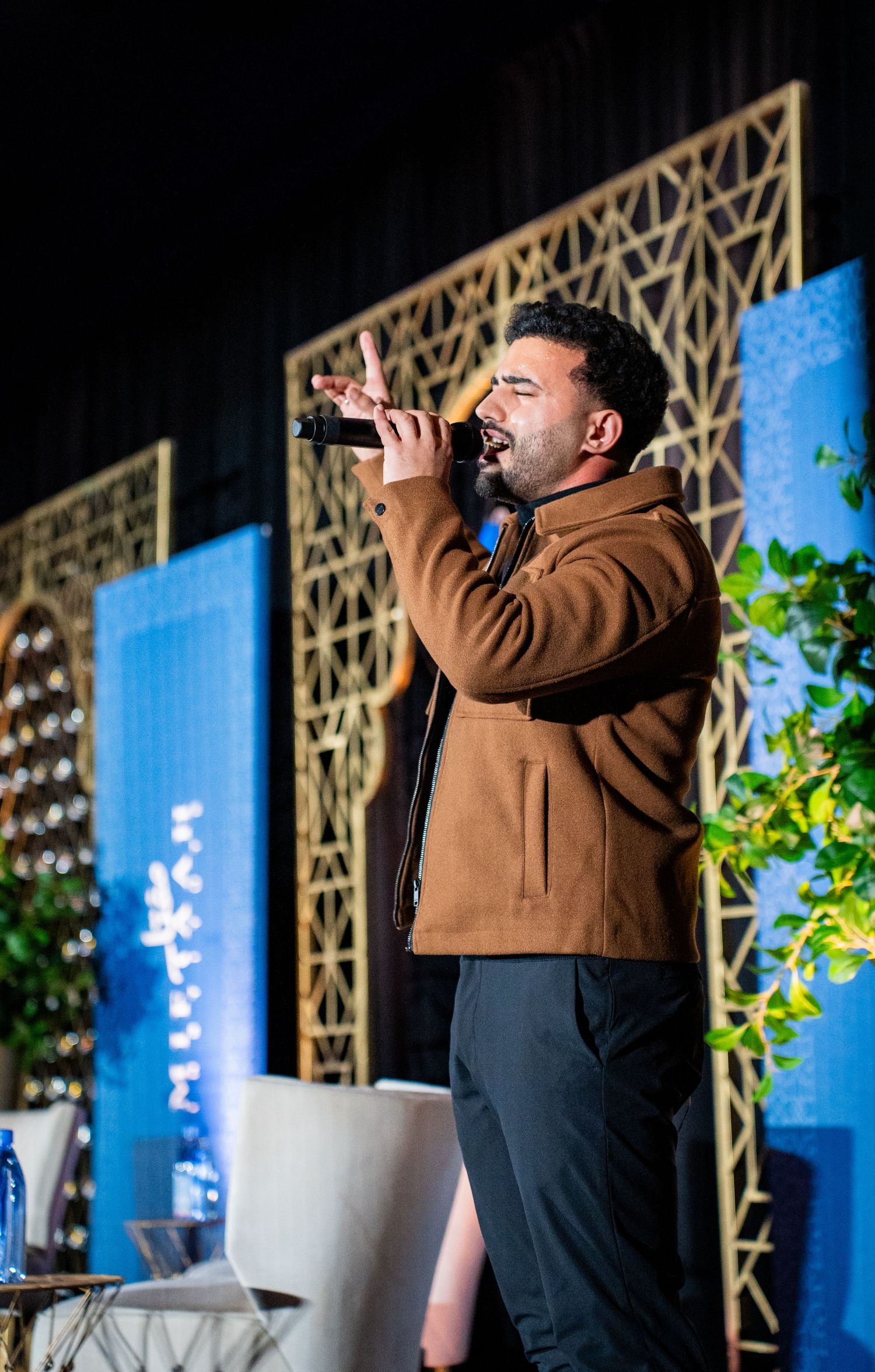 Presenter in brown jacket speaking on stage with geometric backdrop design.