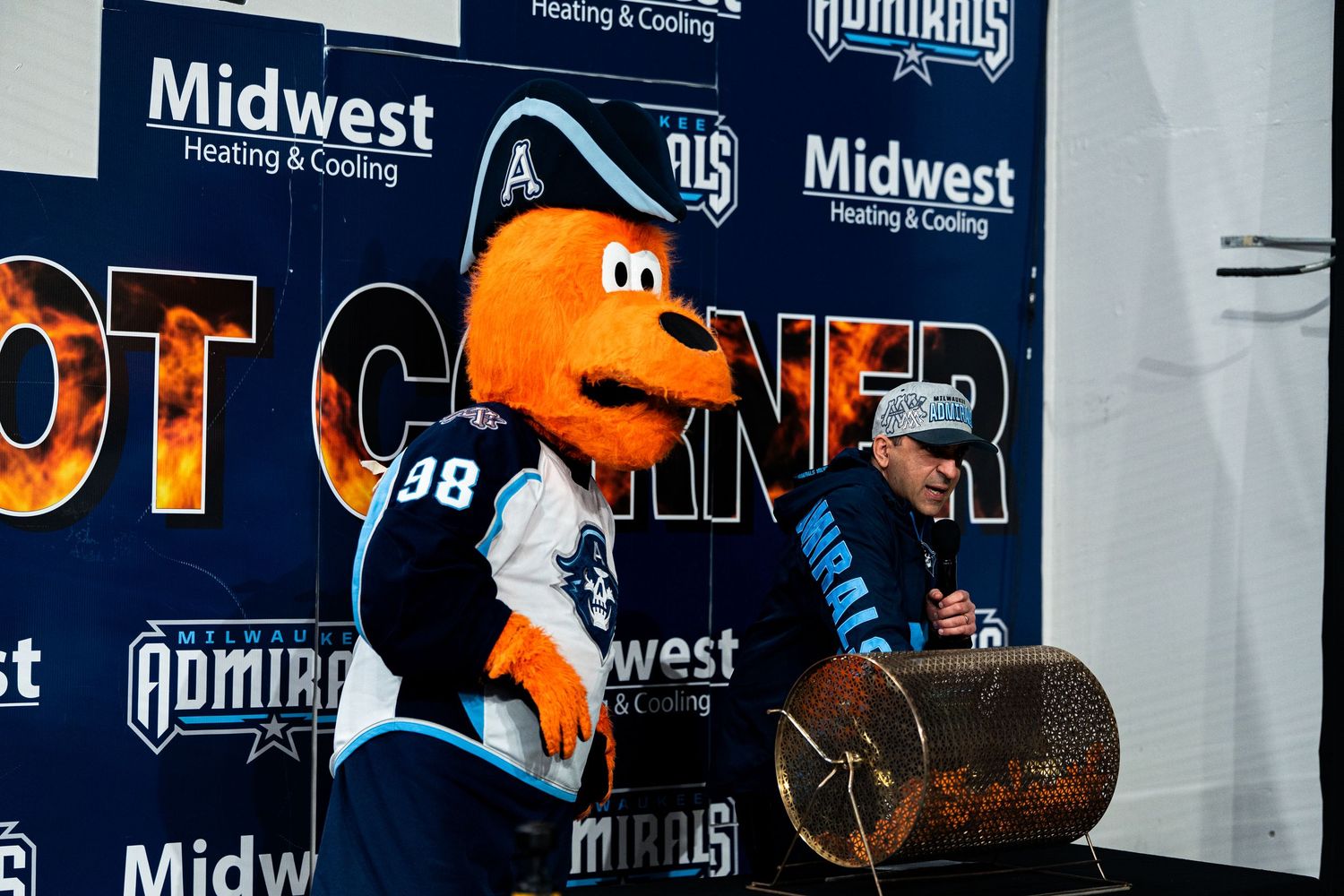 A large orange mascot character interacts with fans at a sports event with Midwest sponsorship banners in the background.