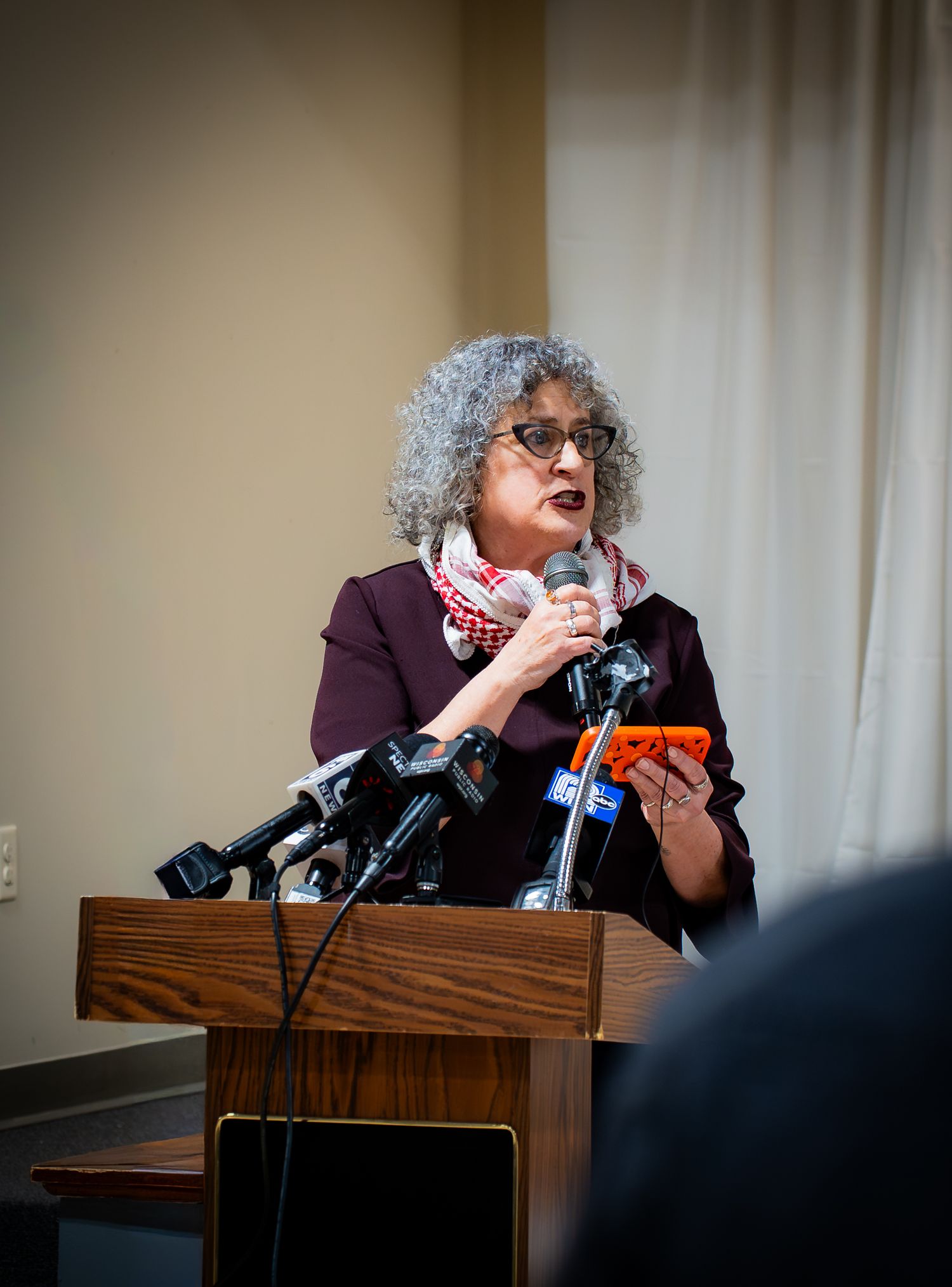 Woman with gray curly hair speaks at podium with microphones, holding phone, wearing burgundy blazer and scarf.