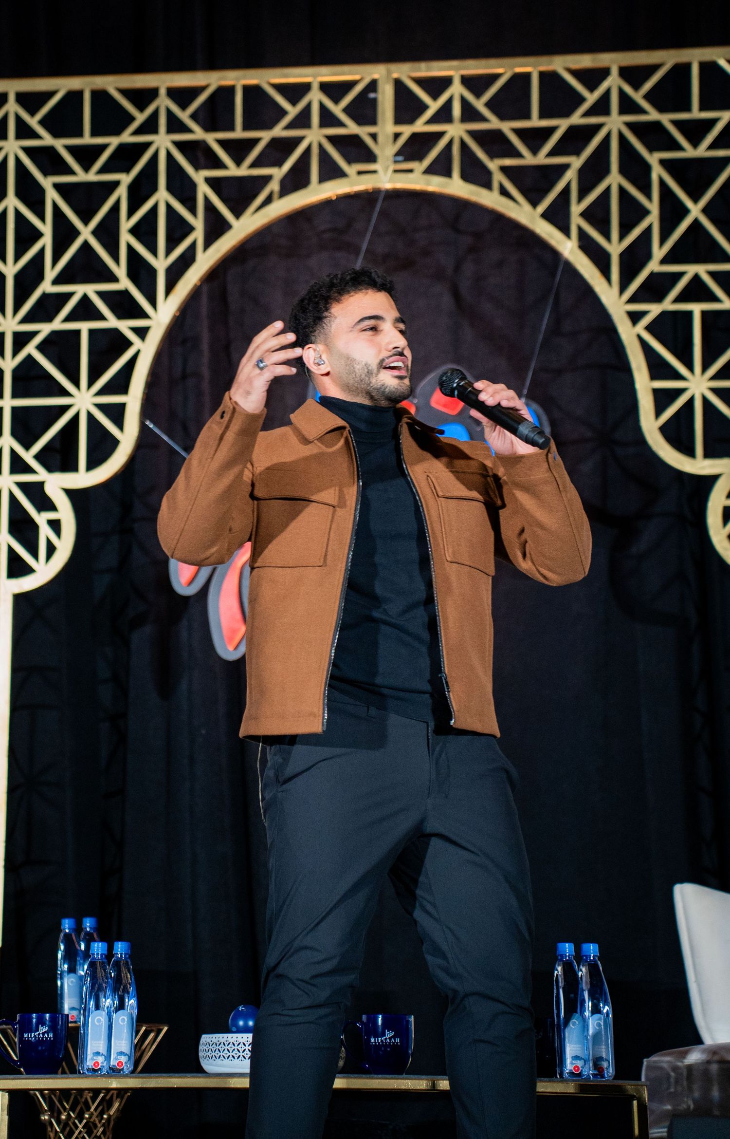 A performer in a brown jacket and dark pants sings on stage with a gold geometric backdrop and water bottles nearby.